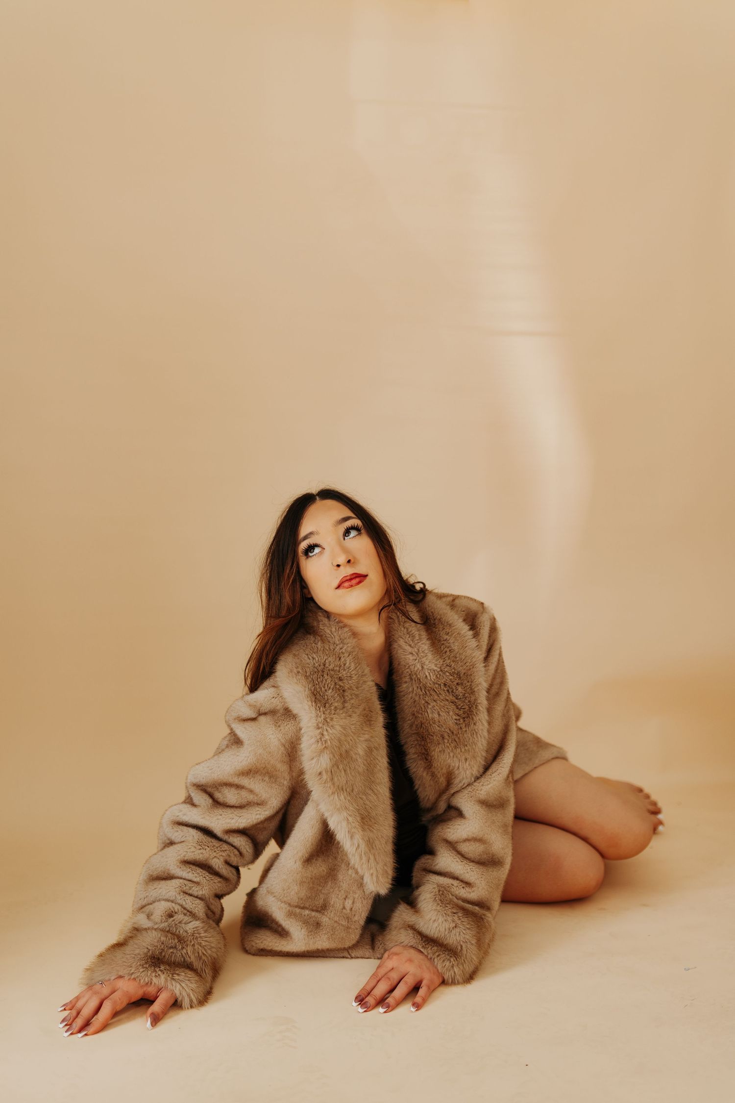 Fashion model in beige fur coat poses artistically on neutral backdrop in minimalist studio setting.