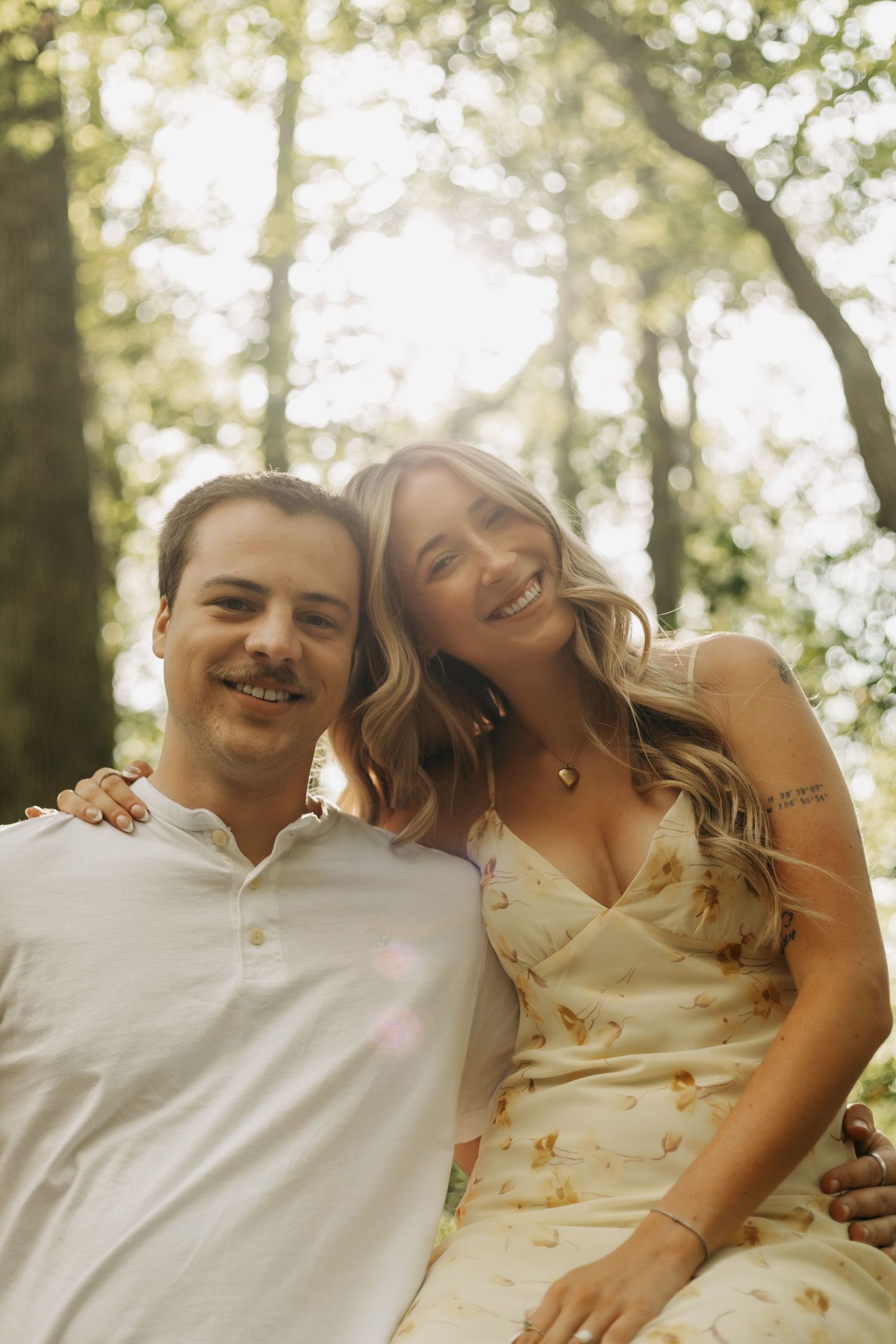 A smiling couple poses together outdoors in a sunlit forest, woman in floral dress with arm around man in white polo.