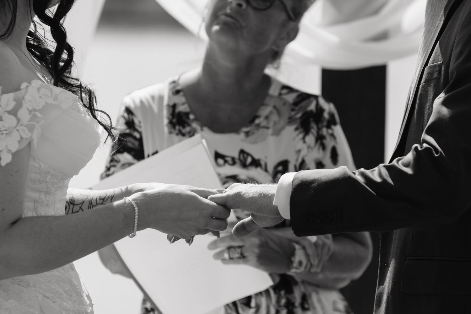 Black and white wedding ceremony photo of couple exchanging rings with officiant in background.