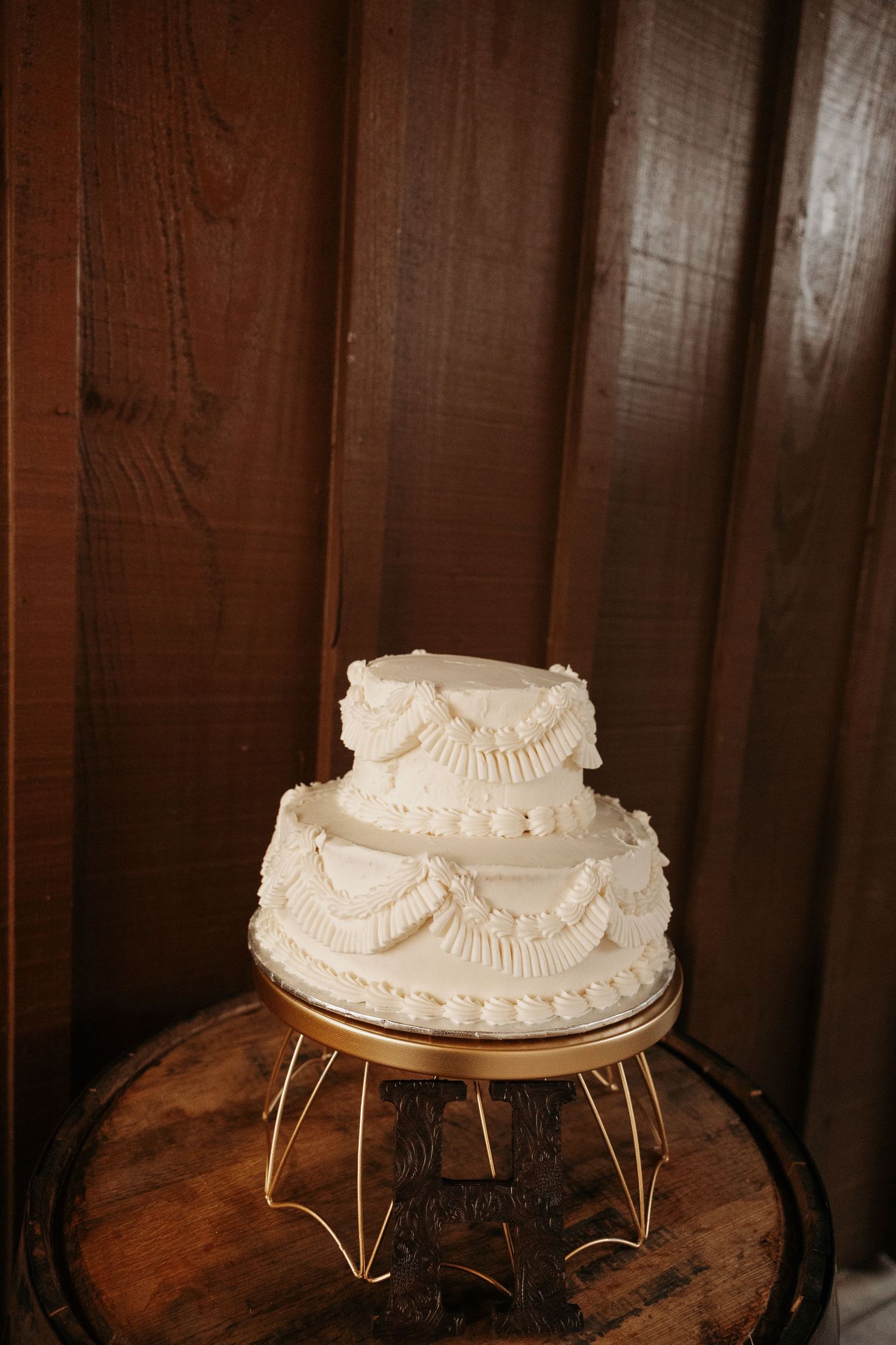 Two-tier white buttercream wedding cake on gold stand against rustic wood wall.