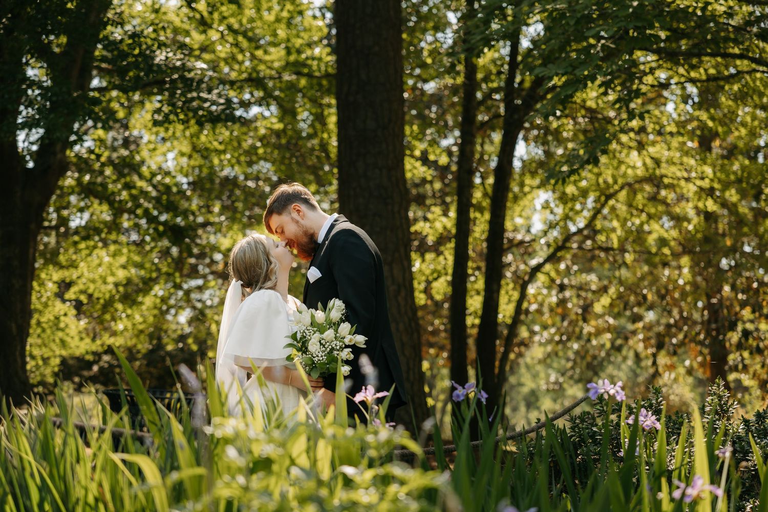 Bride and groom share a romantic kiss surrounded by wildflowers and tall trees in a sunlit garden.