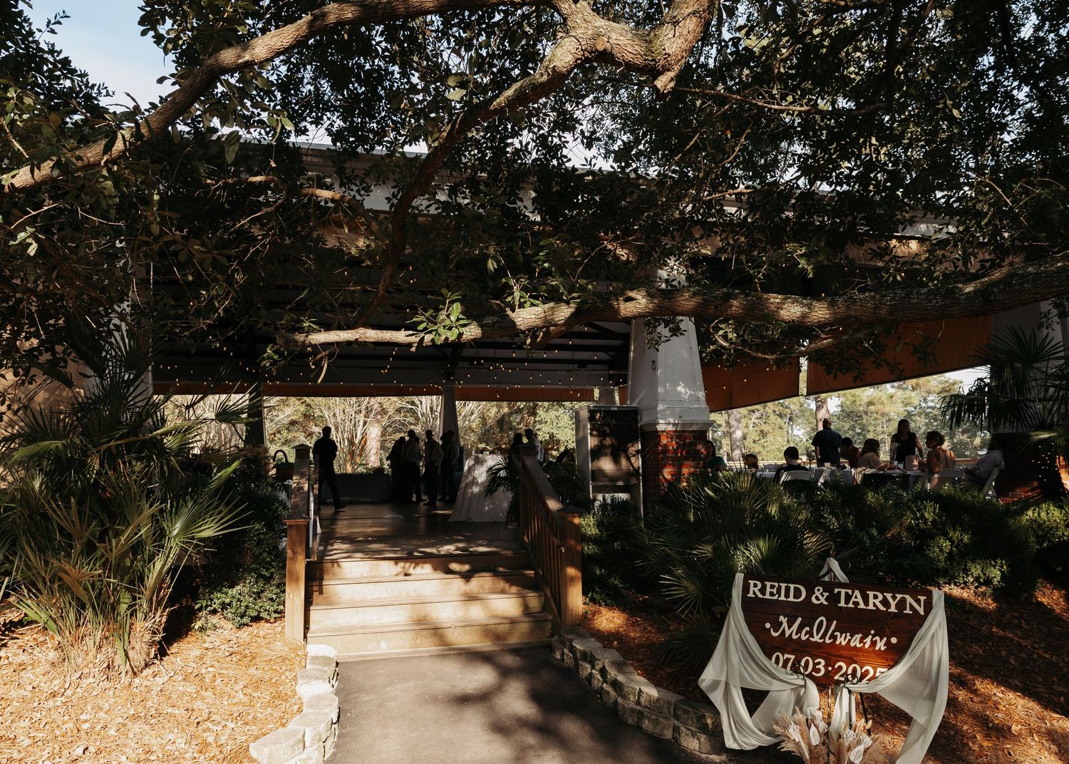 Outdoor wedding ceremony venue with rustic wooden pergola, lush trees, and a welcome sign reading Reid and Taryn.