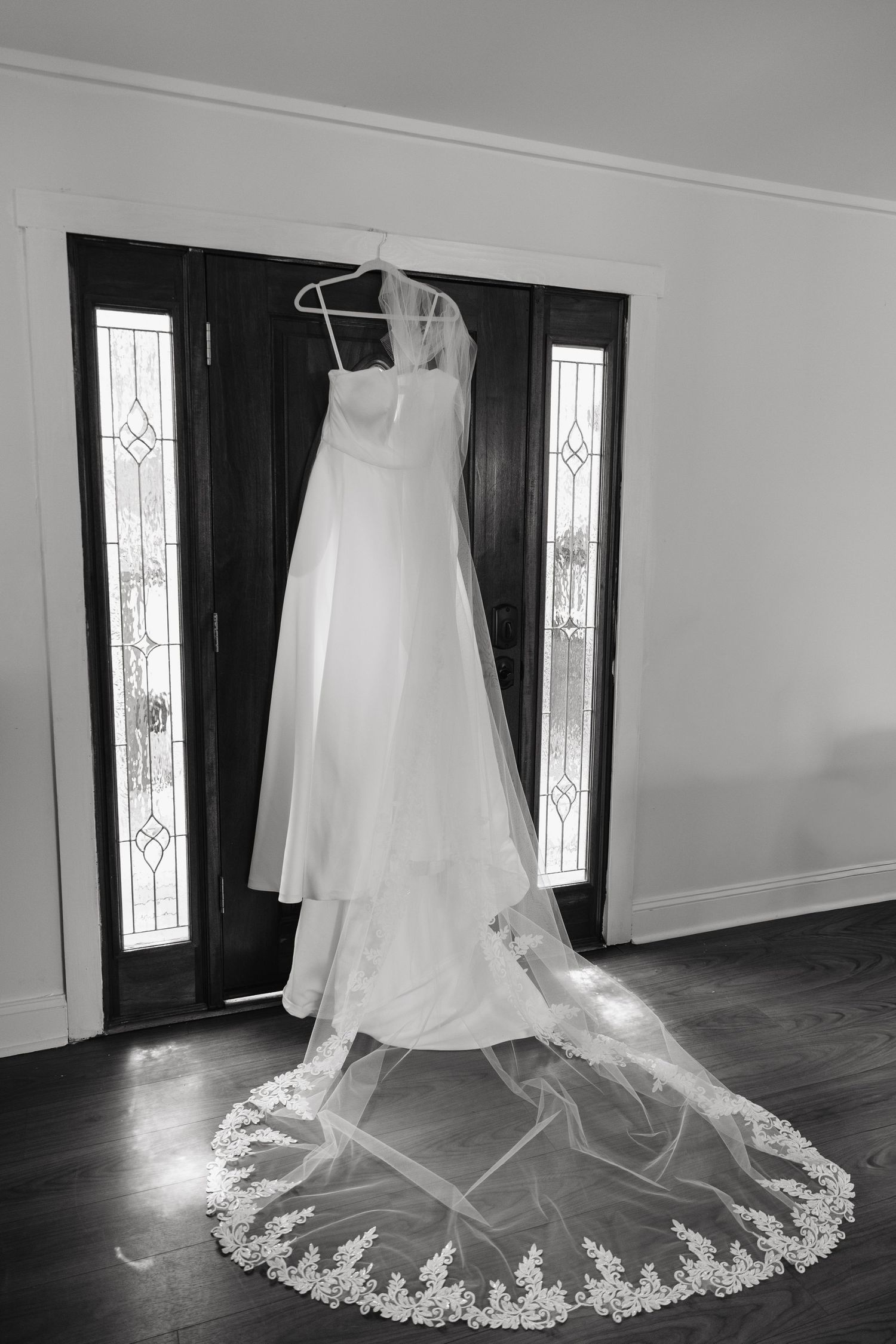 Black and white photo of an elegant wedding dress with lace-trimmed cathedral veil hanging near a door.