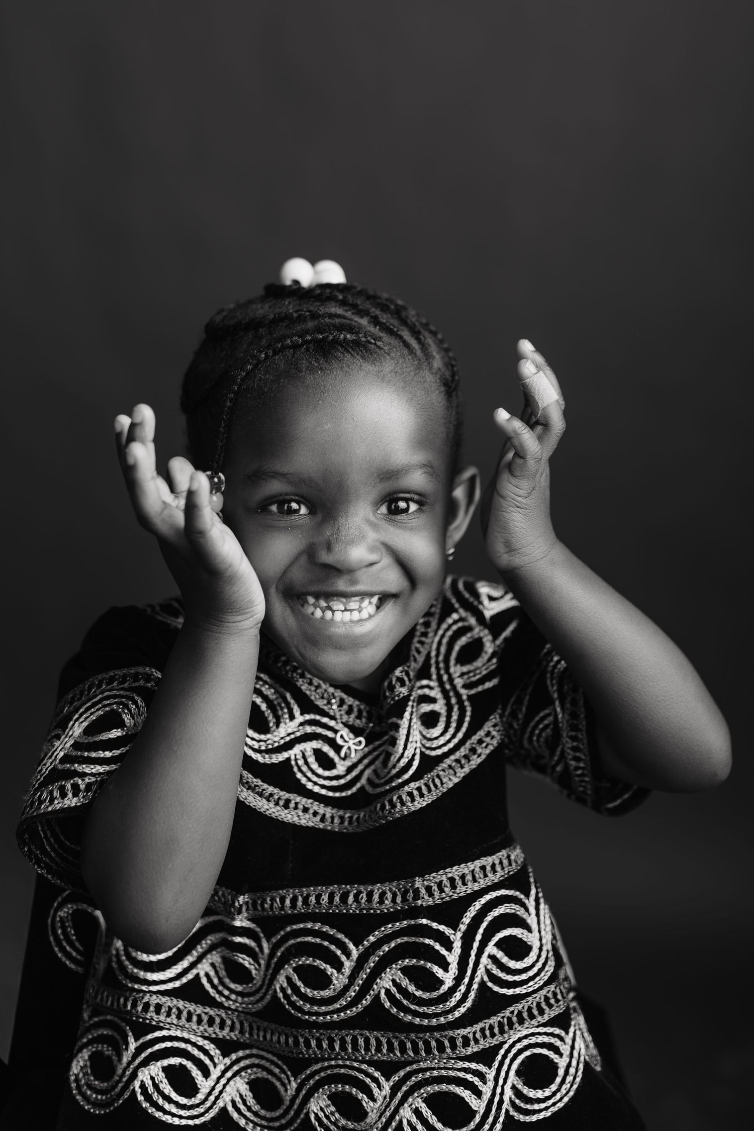Black and white portrait of a joyful young African girl in traditional embroidered dress, hands raised, smiling brightly.