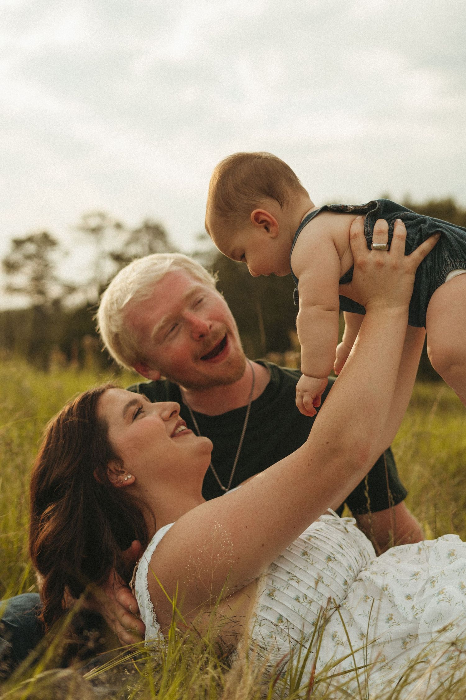 A joyful family sits in a grassy field, parents laughing as they lift their happy baby in the air.