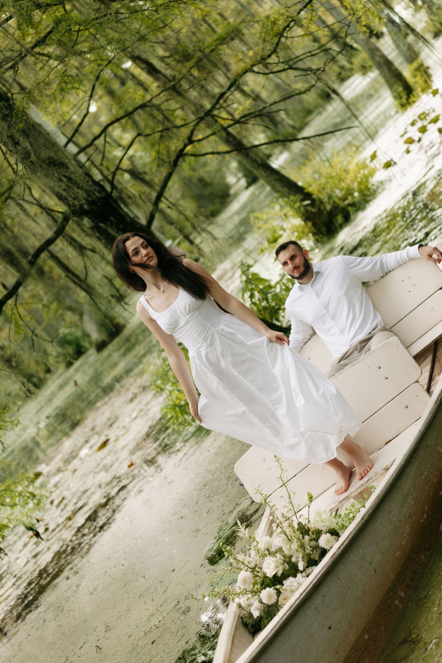 Smiling bride in white dress standing on a flower-adorned boat with groom in lush swamp.