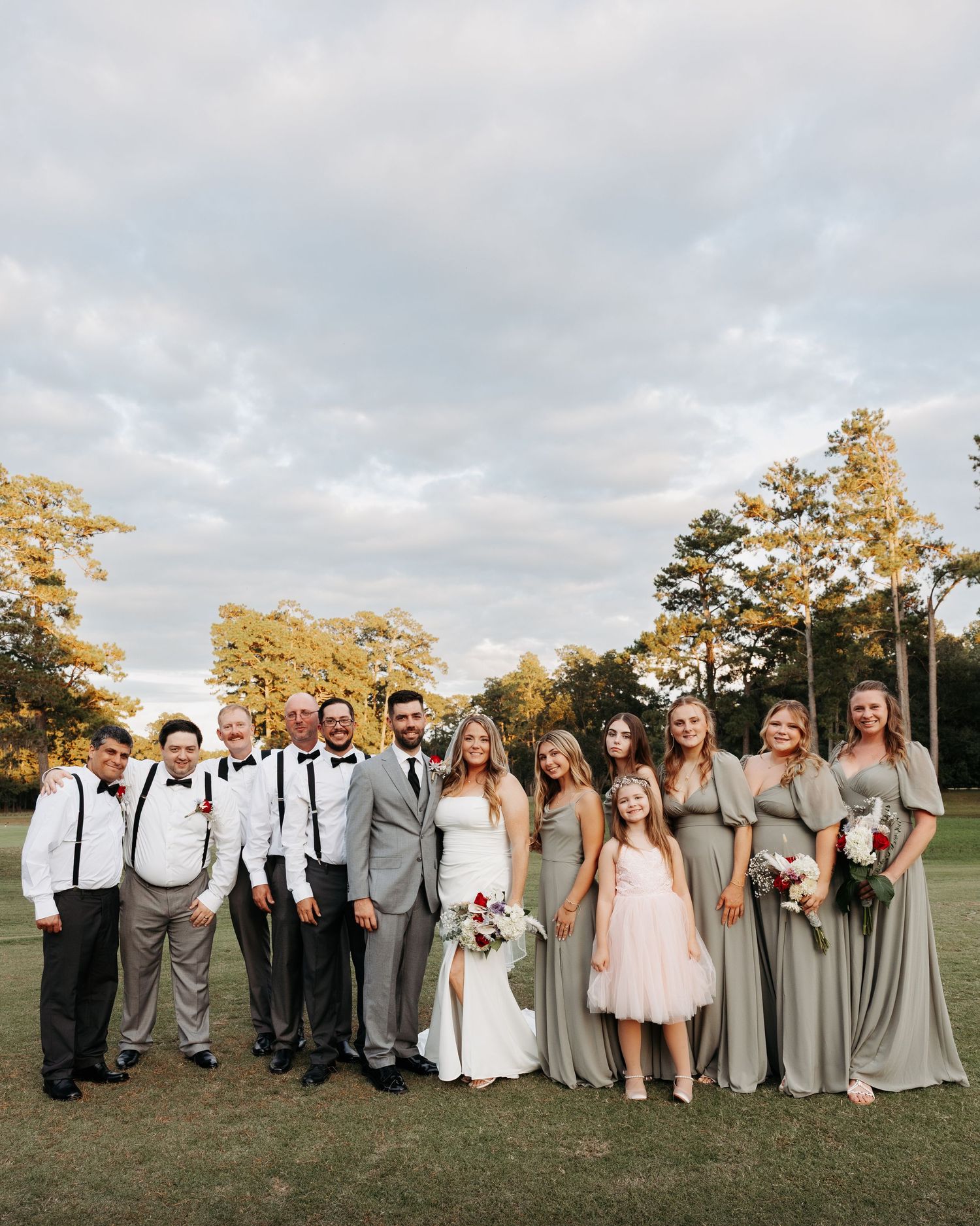 Wedding party in grey and white attire poses together on a grassy field during golden hour.