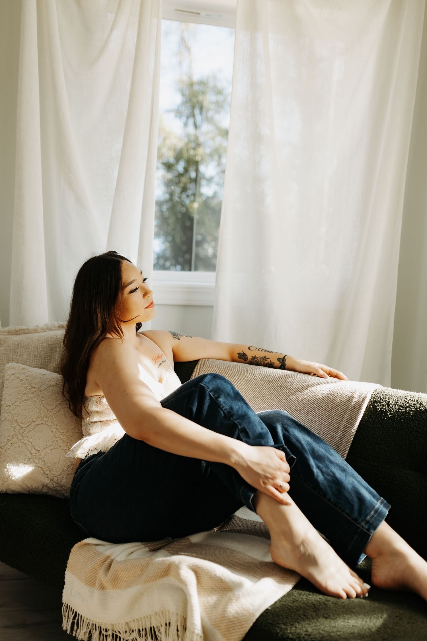 A relaxed person lounges on a couch by sunlit window with white curtains, wearing jeans and a light top.