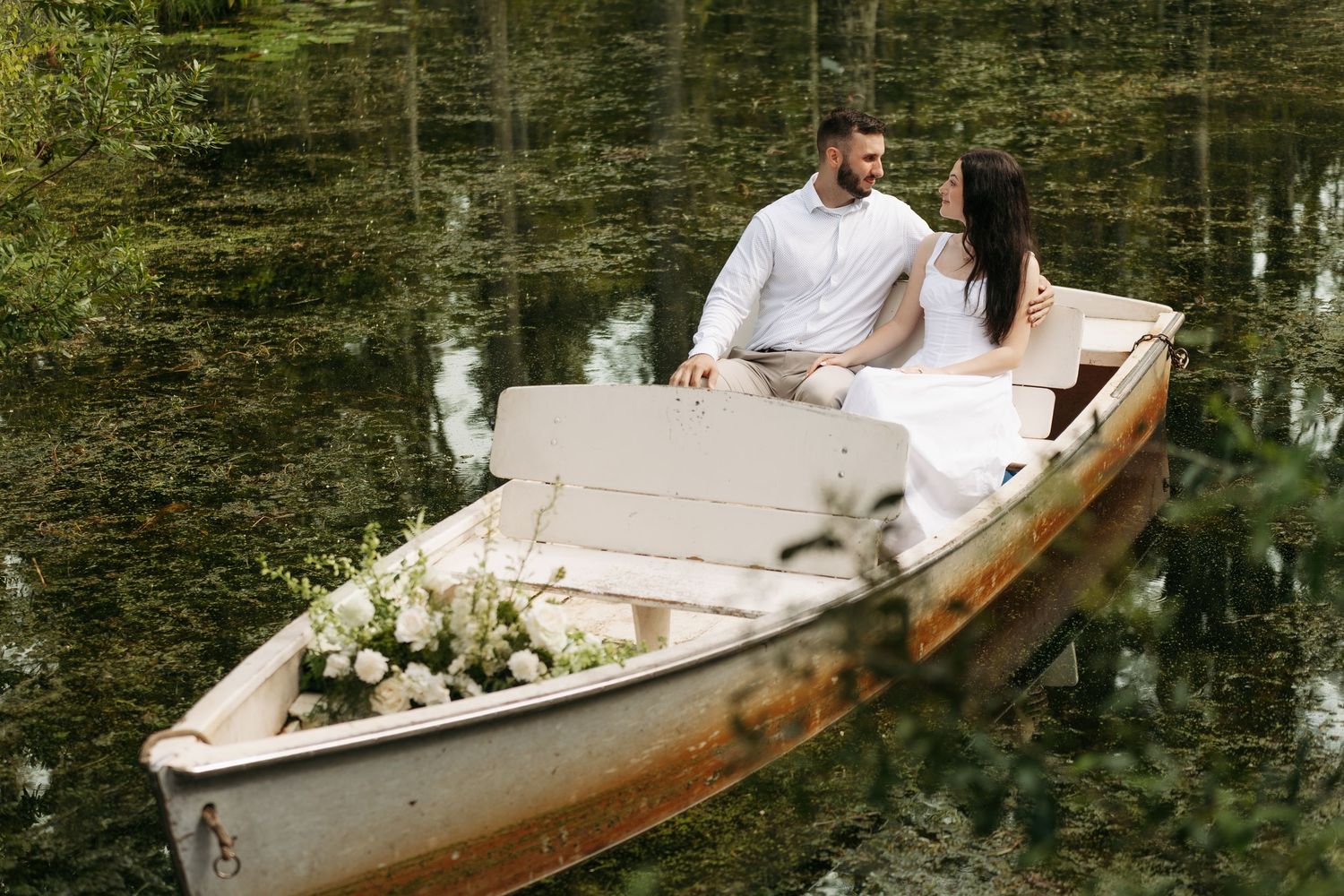 Couple sitting romantically in a flower-adorned wooden boat on a calm, tree-reflected lake.