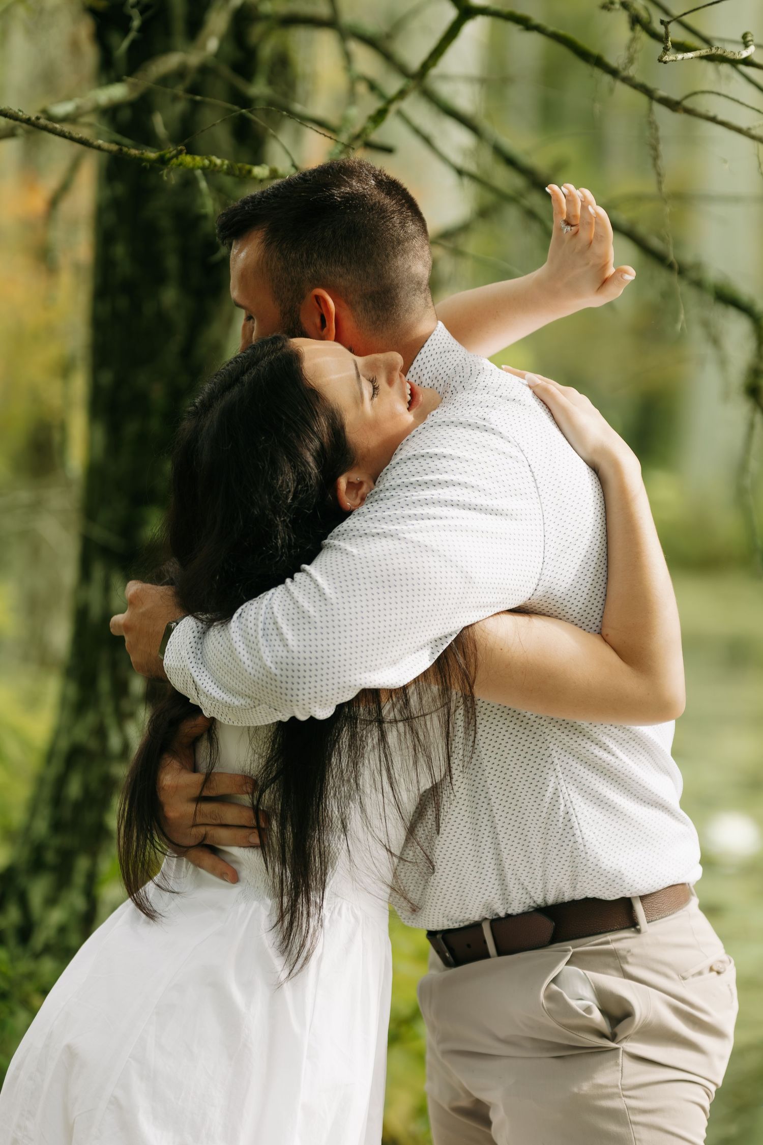 Romantic couple embracing and kissing outdoors under green trees in a lush garden setting.