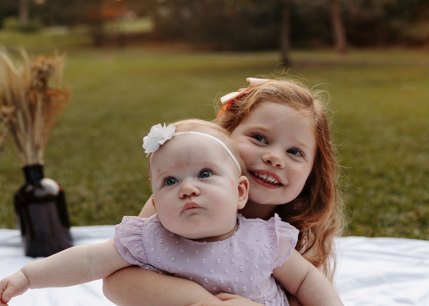 A smiling young girl hugs a baby in a pink dress on a white blanket outdoors in a grassy park setting.
