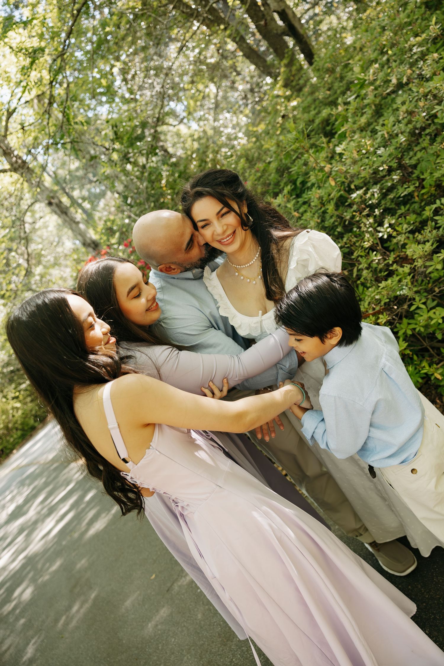 A joyful family of five shares a warm embrace outdoors, surrounded by lush green trees on a sunny day.