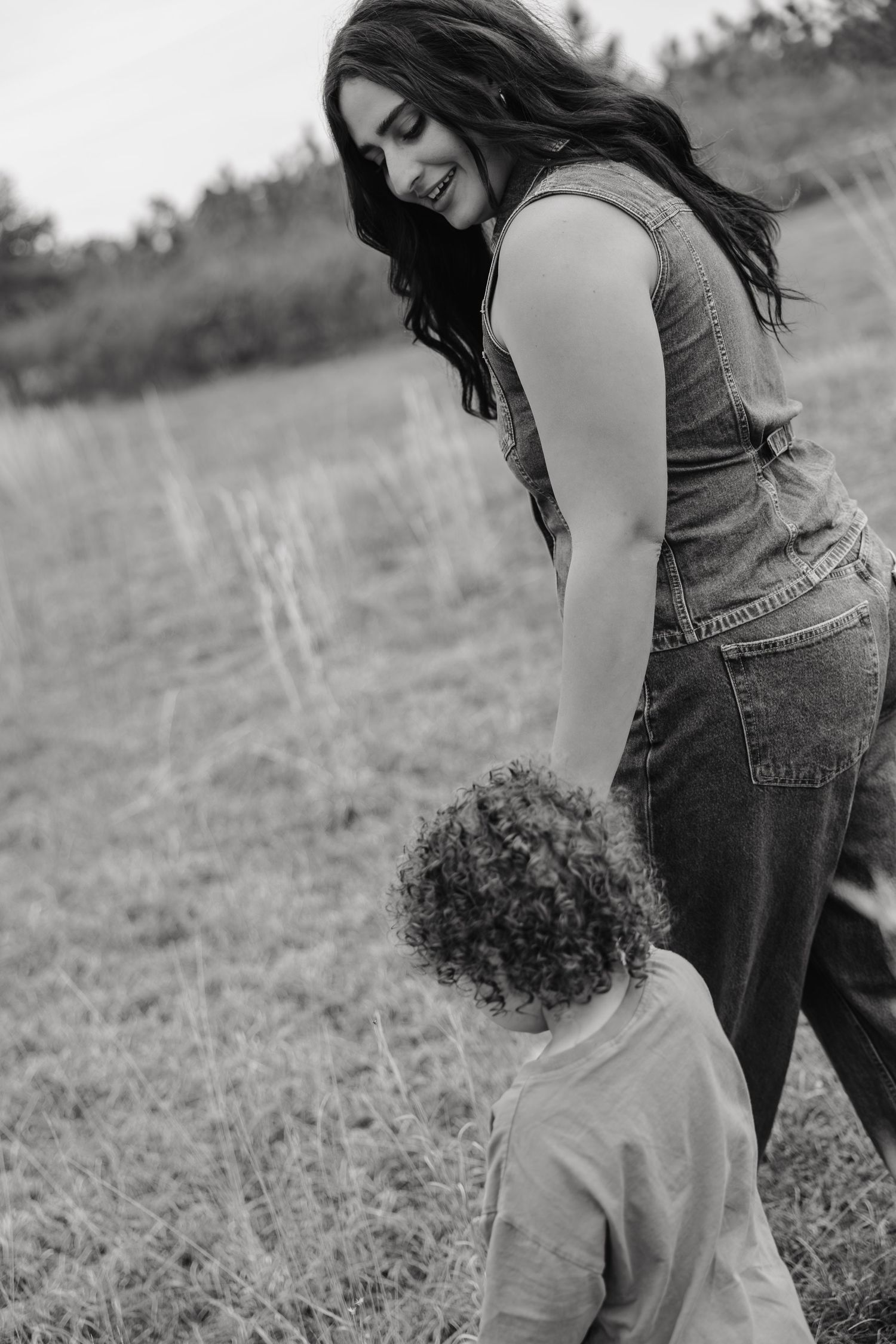 Black and white photo of a smiling mother holding hands with her curly-haired toddler in an open field.