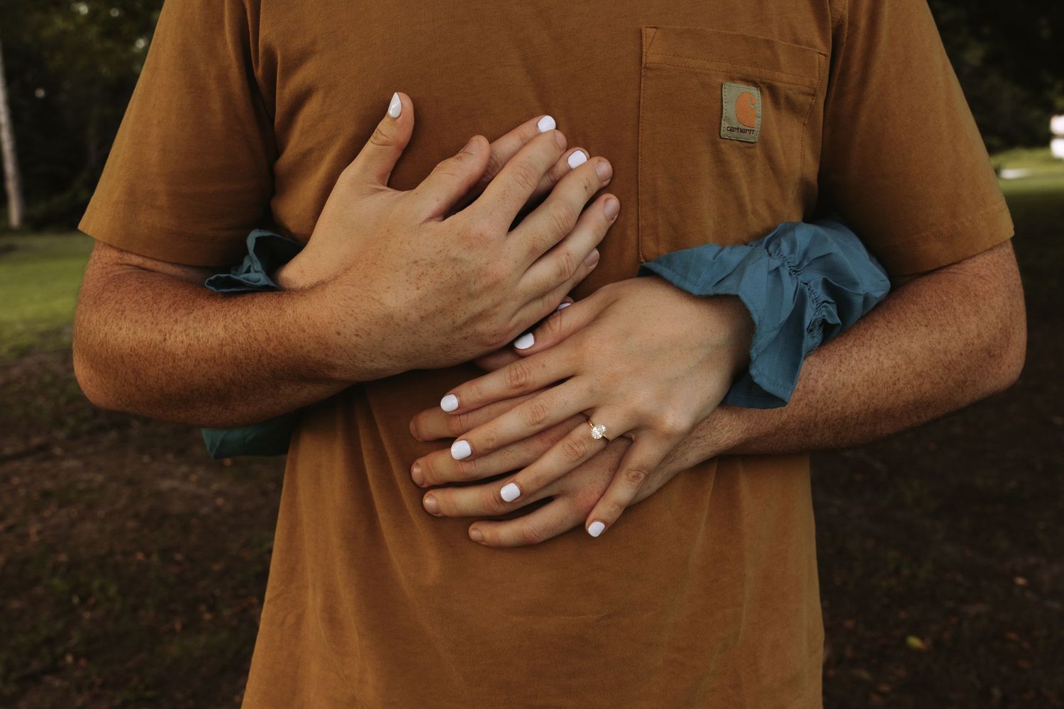 Hands crossed over a brown t-shirt showing engagement or wedding rings in an outdoor setting at dusk.