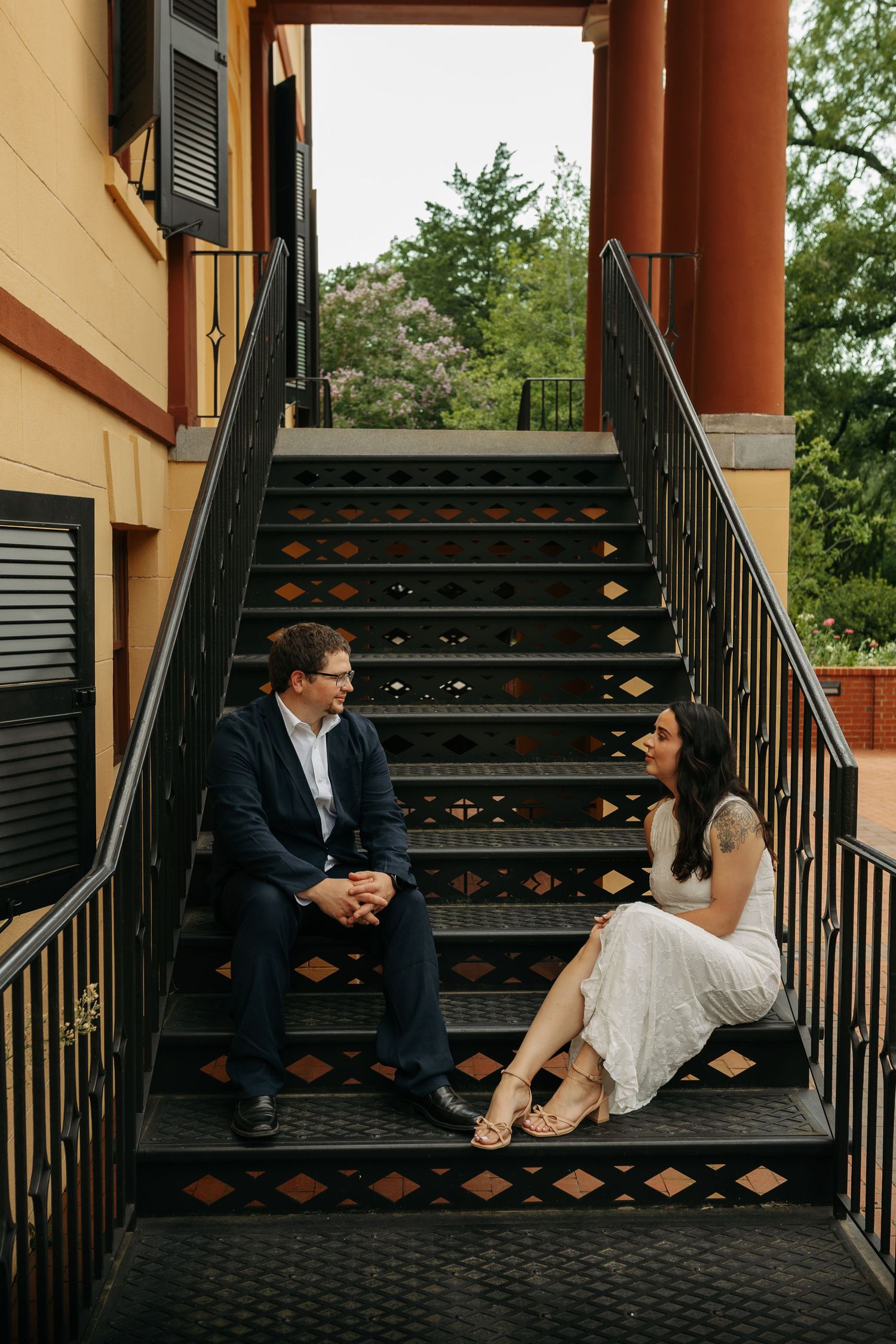 Couple sitting on decorative black metal stairs of a yellow building, sharing a romantic conversation outdoors.