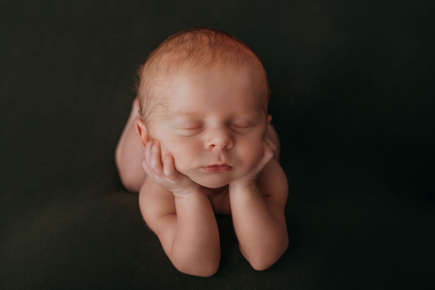 Peaceful newborn baby sleeping with hands under chin on dark green backdrop in professional studio photography.