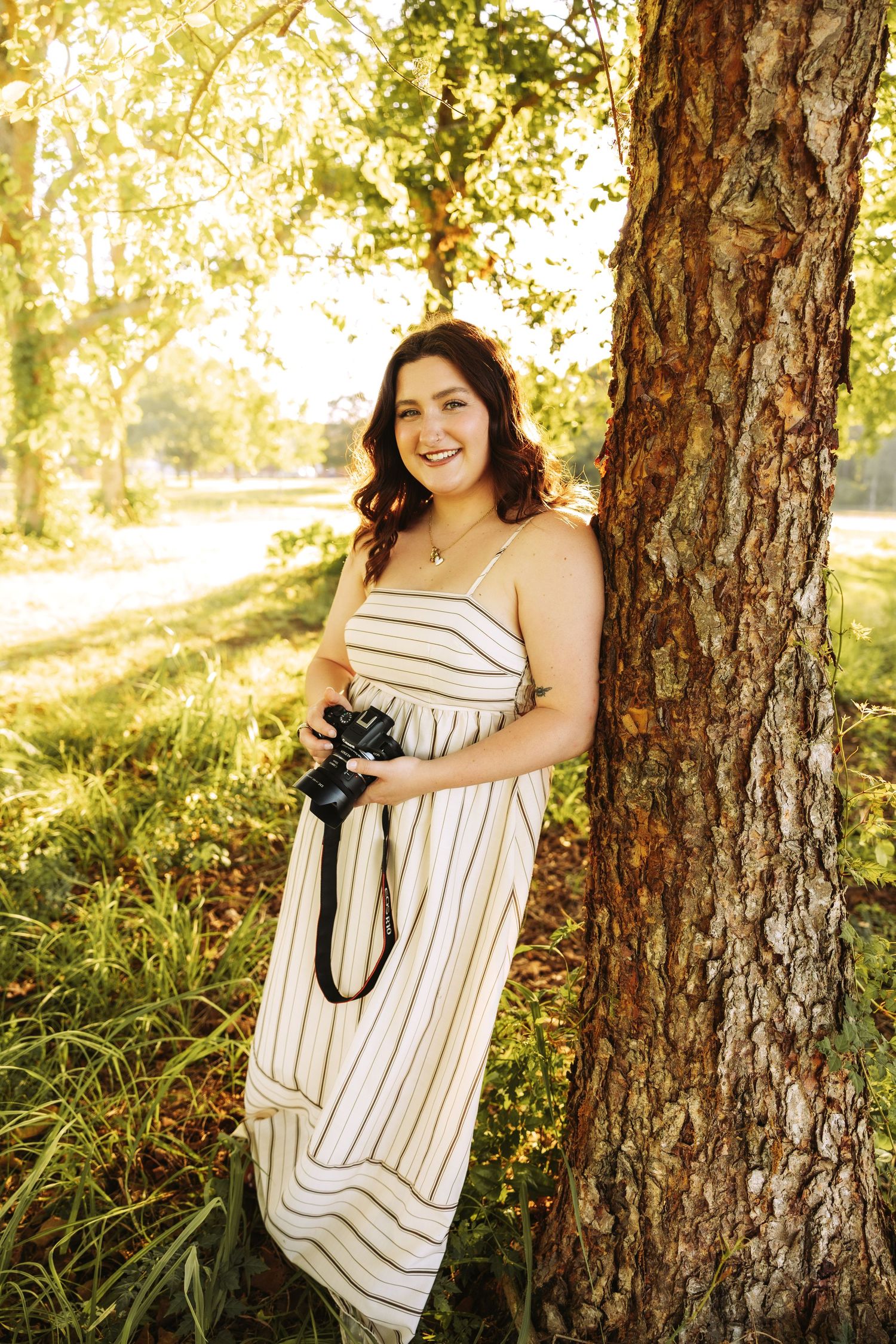 Young woman in striped sundress holding camera, leaning against tree in sunlit park, smiling warmly.