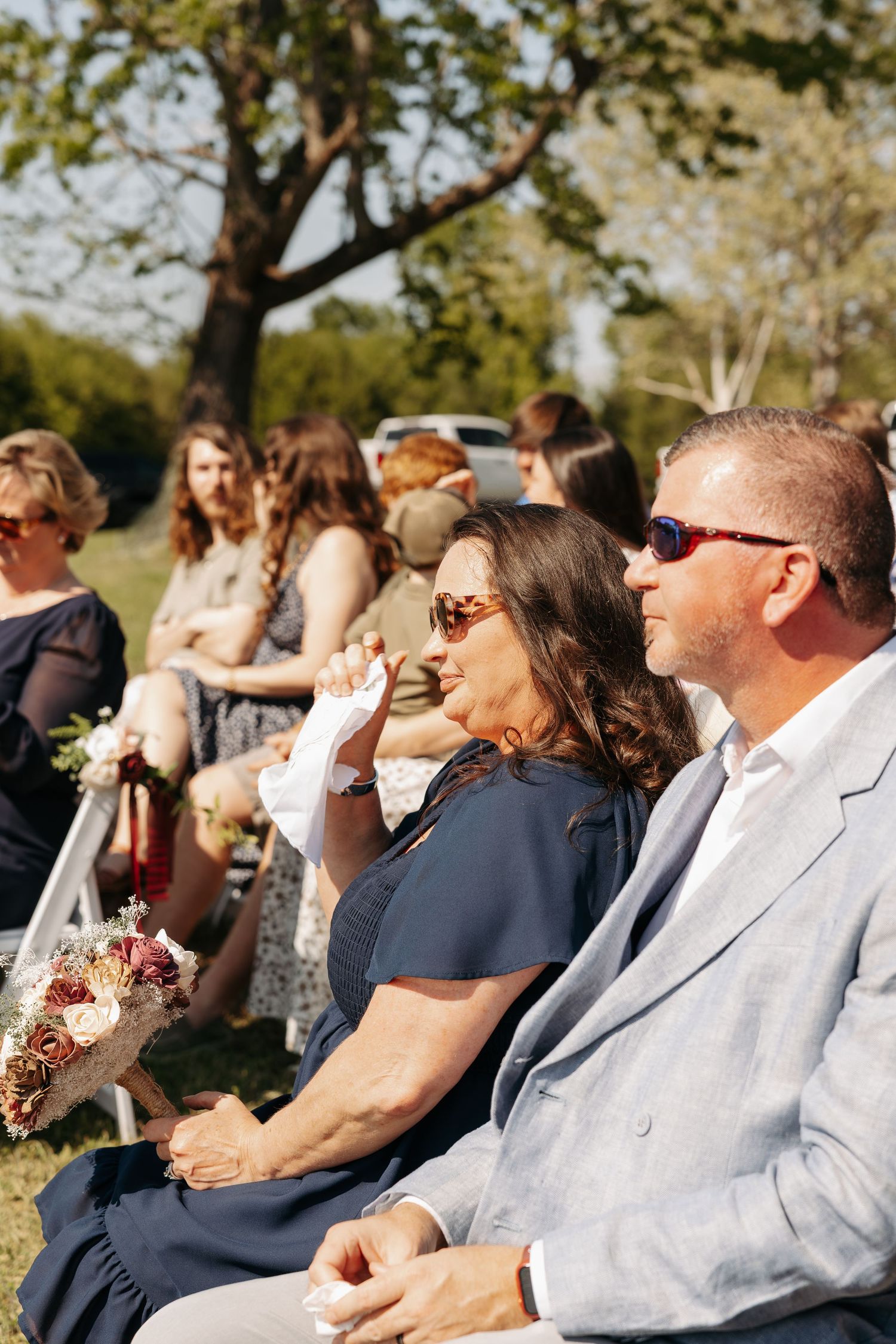 Wedding guests seated outdoors in sunlight, woman in navy dress holding bouquet beside man in light gray suit wearing sunglasses.