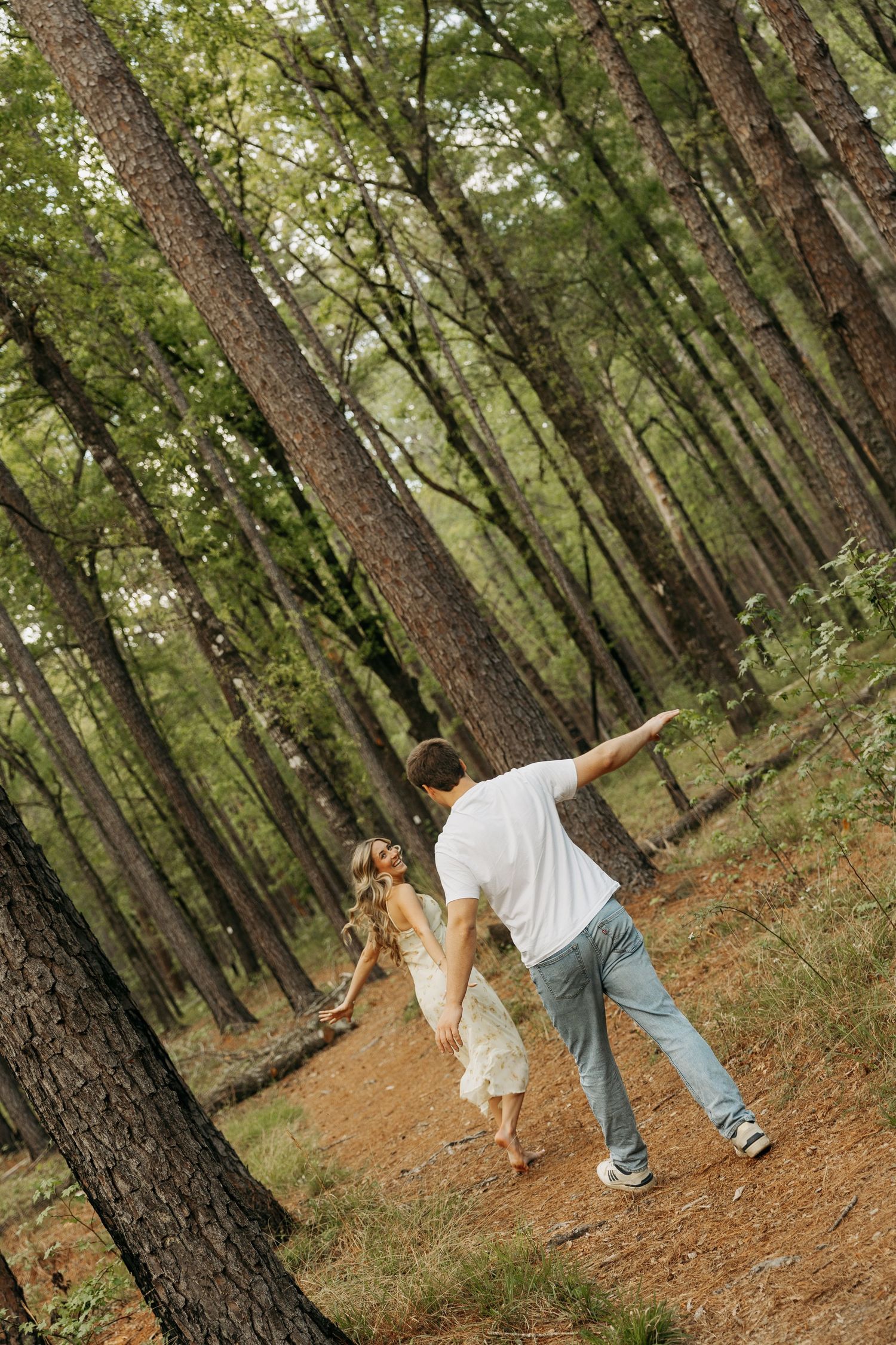 Couple dancing playfully in a pine forest, man in white shirt and jeans spinning woman in white dress on a dirt path.