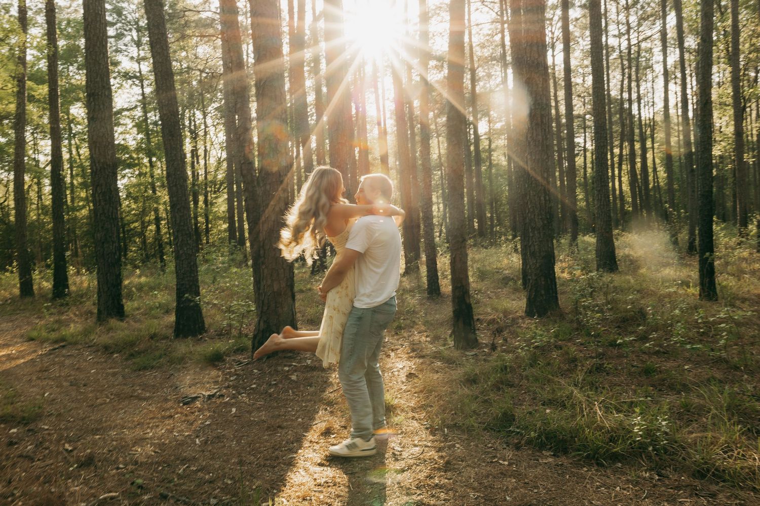 A couple embraces joyfully in a sunlit forest, with golden rays streaming through tall pine trees.