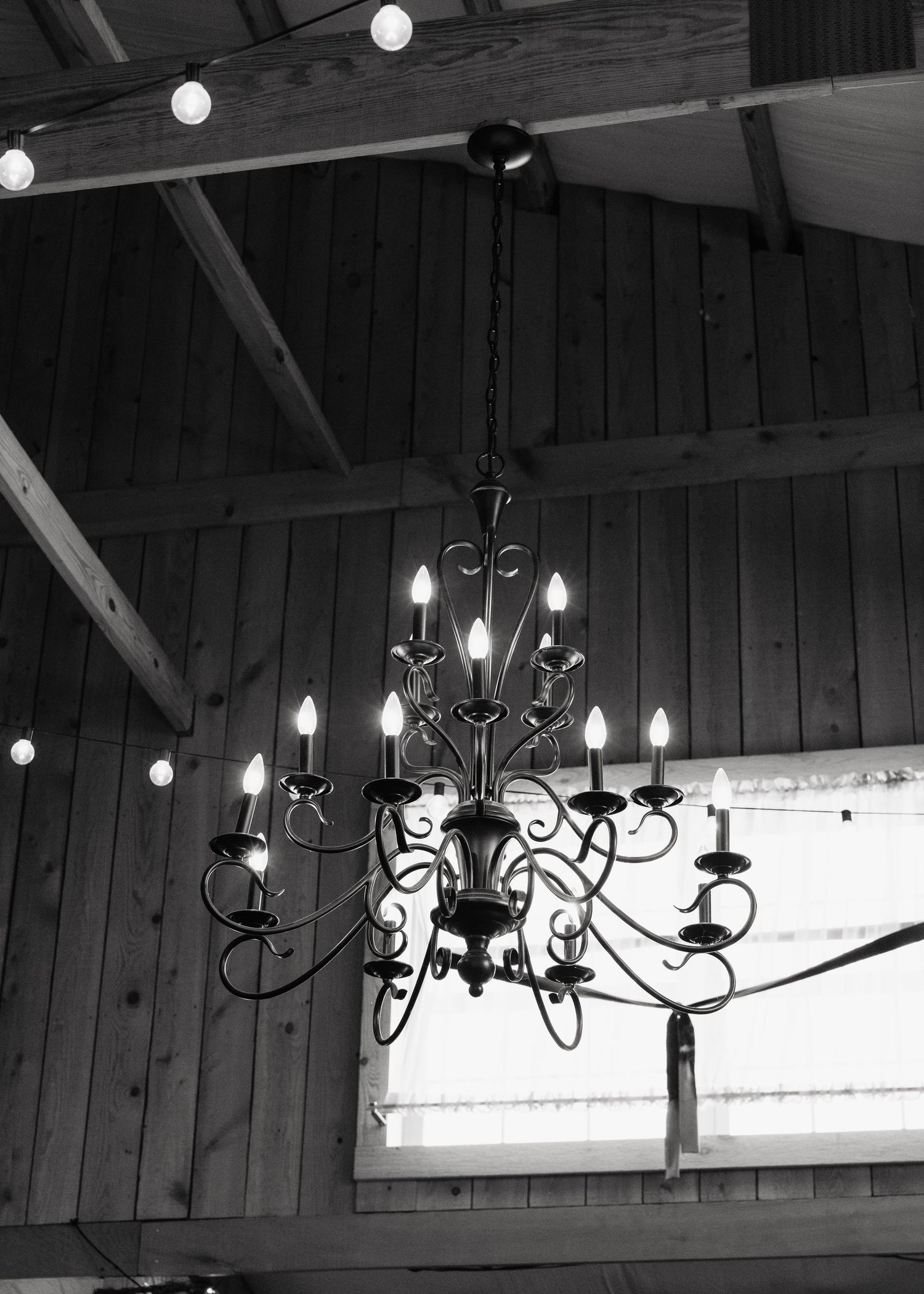 Black and white photo of an elegant wrought iron chandelier with candle lights hanging from a rustic wooden ceiling.