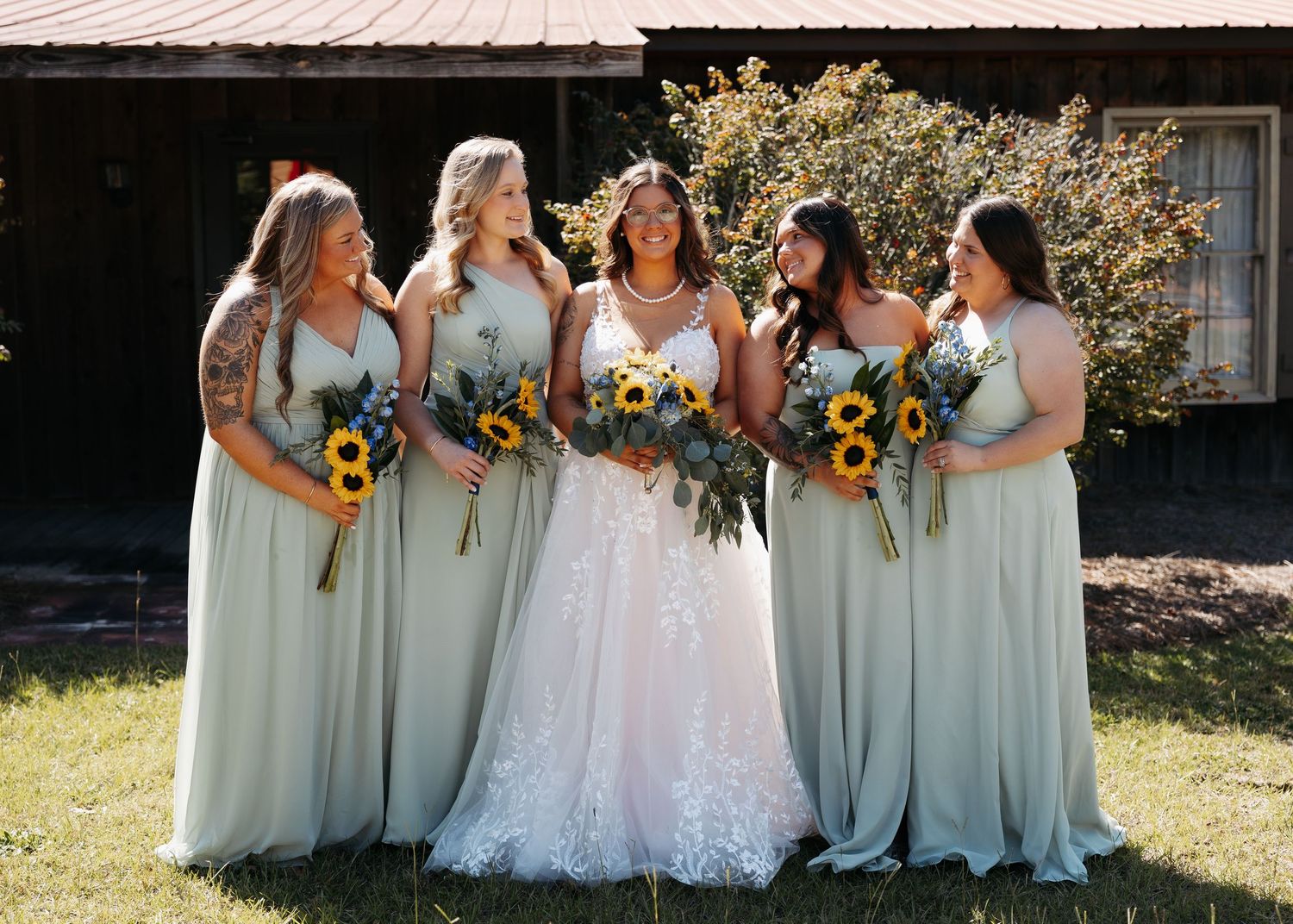 Bride in lace gown with four bridesmaids in sage green dresses holding sunflower bouquets outdoors.
