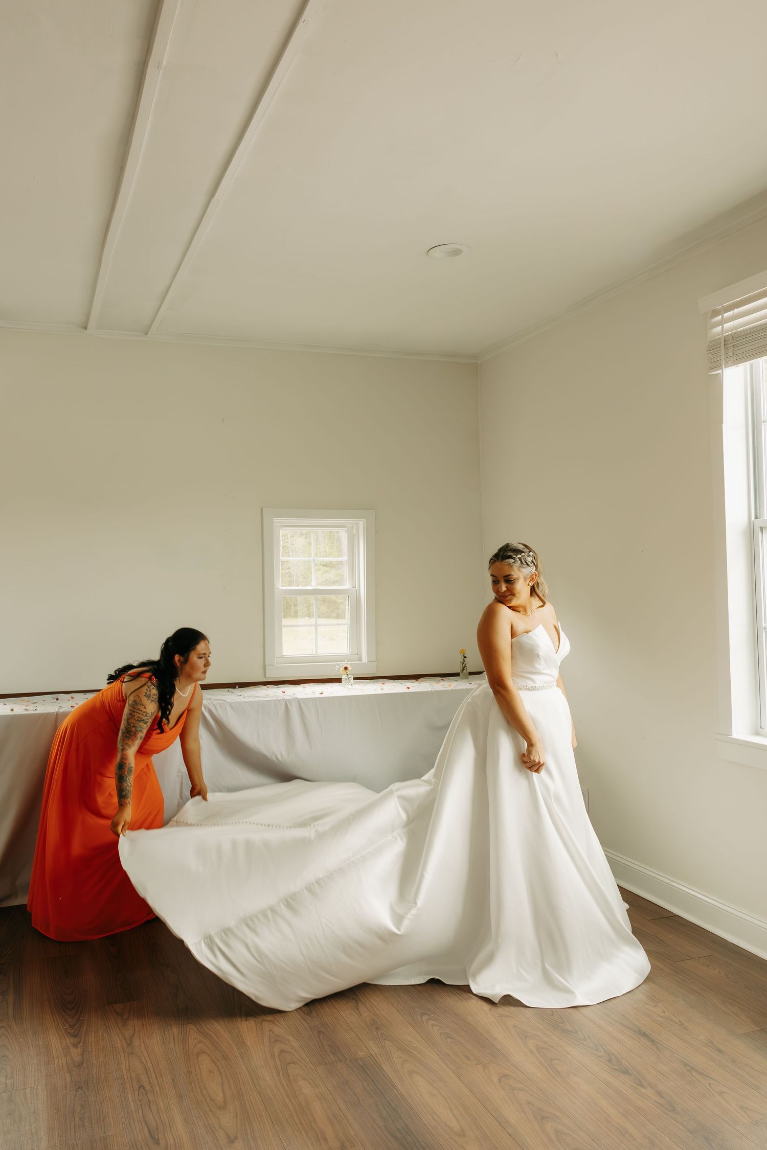 Bridesmaid in orange dress helps bride arrange her flowing white wedding gown train in a bright room.