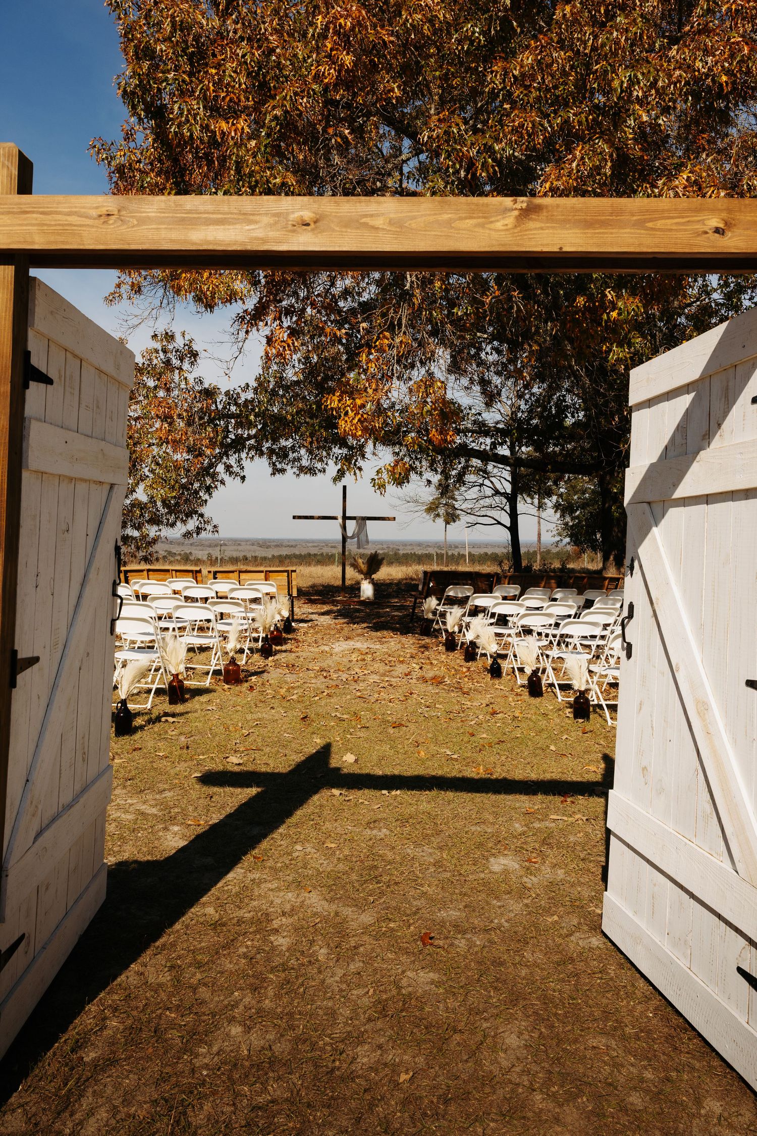 Outdoor wedding ceremony setup with white chairs, wooden cross altar, barn doors, and autumn trees under a clear blue sky.