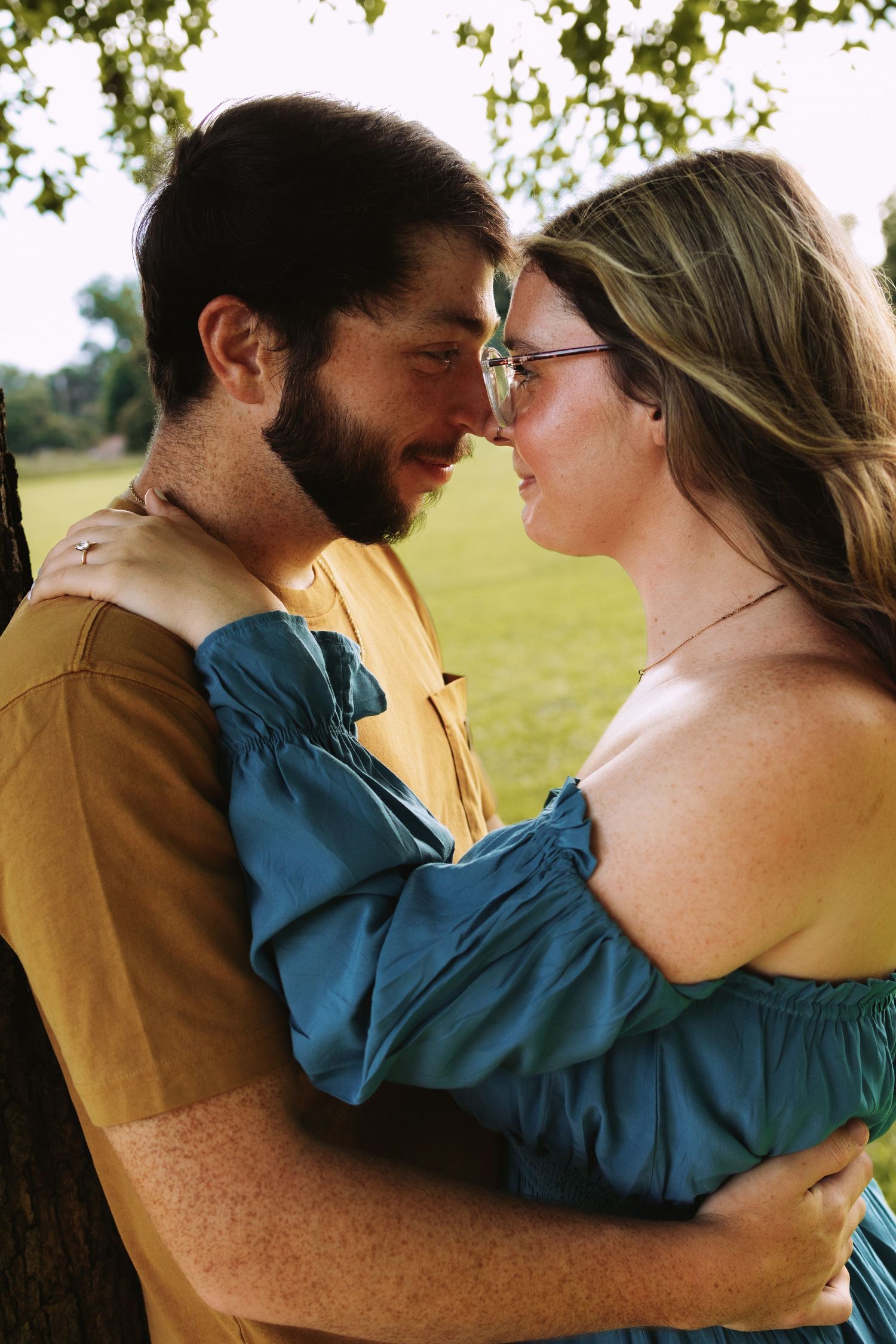 A bearded man and woman in a teal dress share a romantic embrace outdoors near a tree.