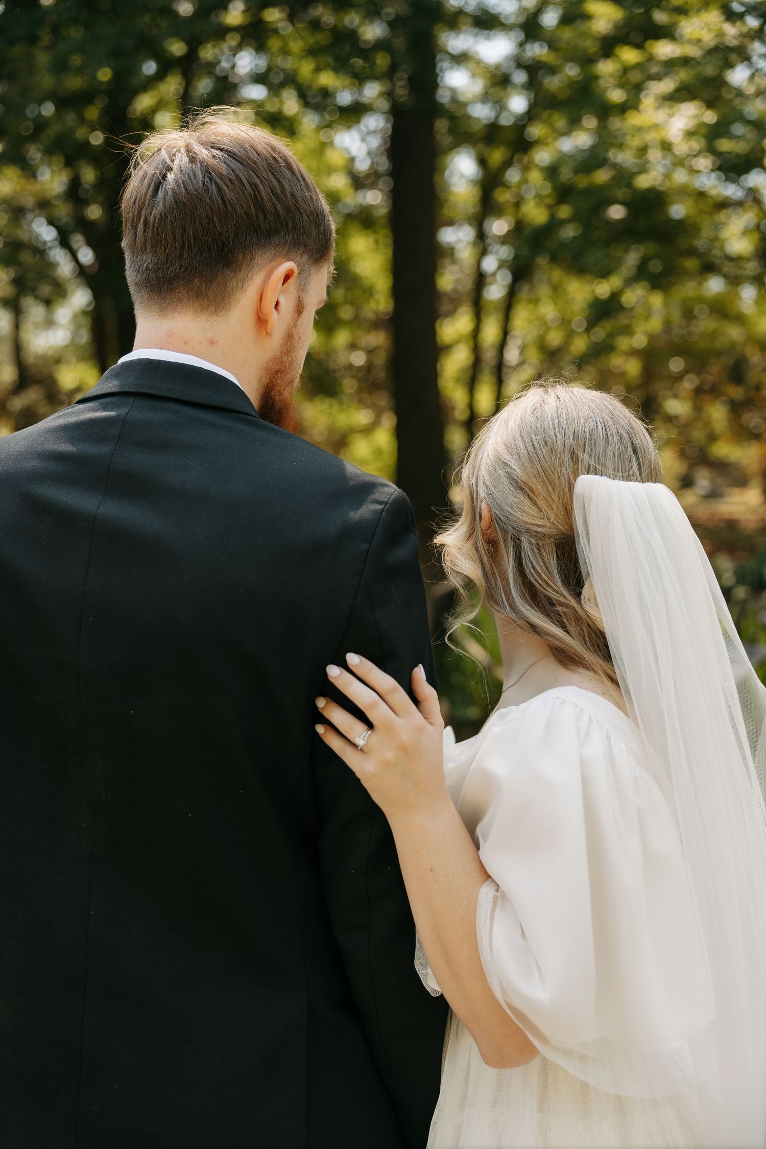 Rear view of bride and groom walking together through a sunlit forest path.