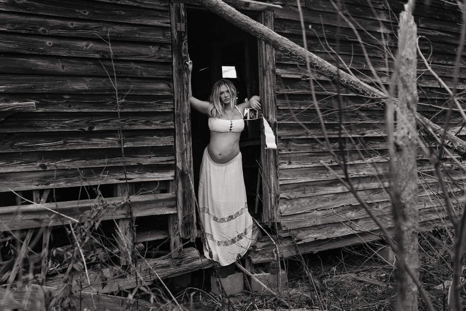 Pregnant woman in white dress standing in doorway of rustic abandoned wooden cabin in black and white photo.