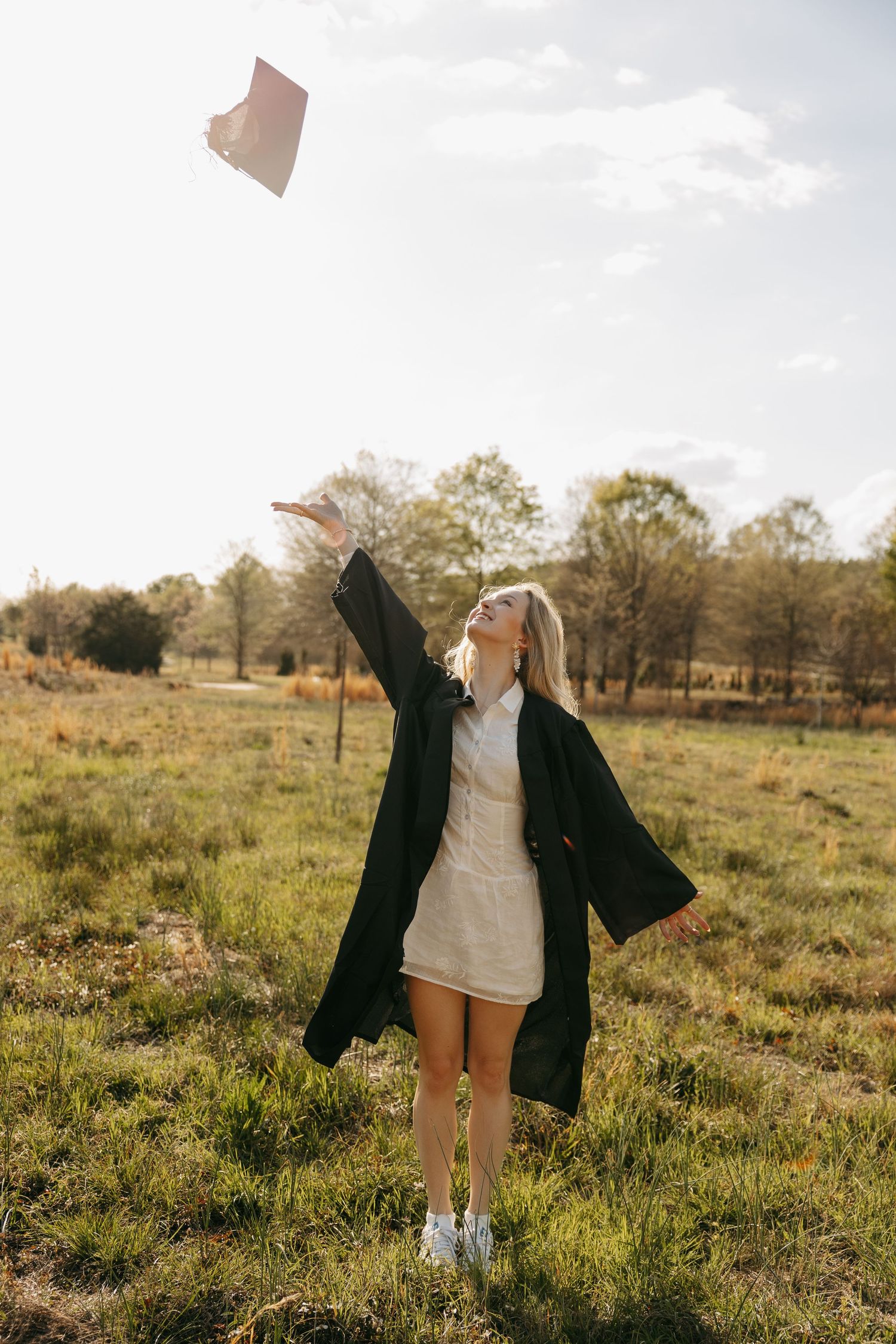 A joyful graduate in black gown tosses her cap in a sunny field, celebrating her academic achievement outdoors.