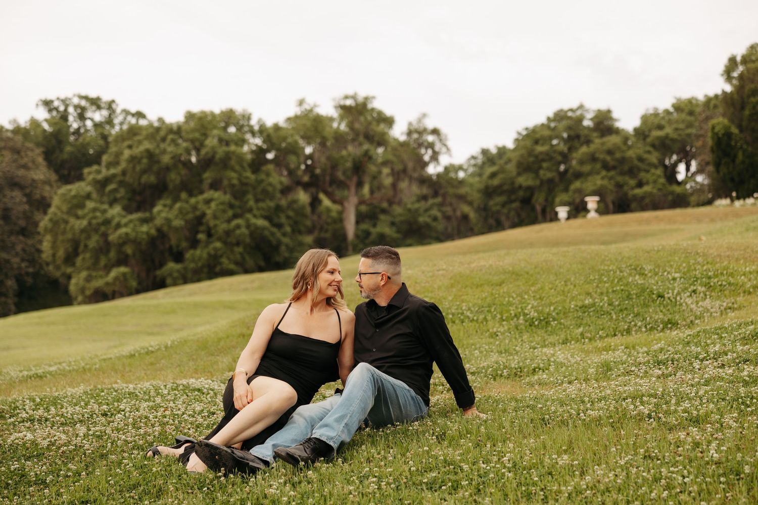 Couple sitting together on a lush green hillside, woman in black dress and man in black shirt, sharing a romantic moment.