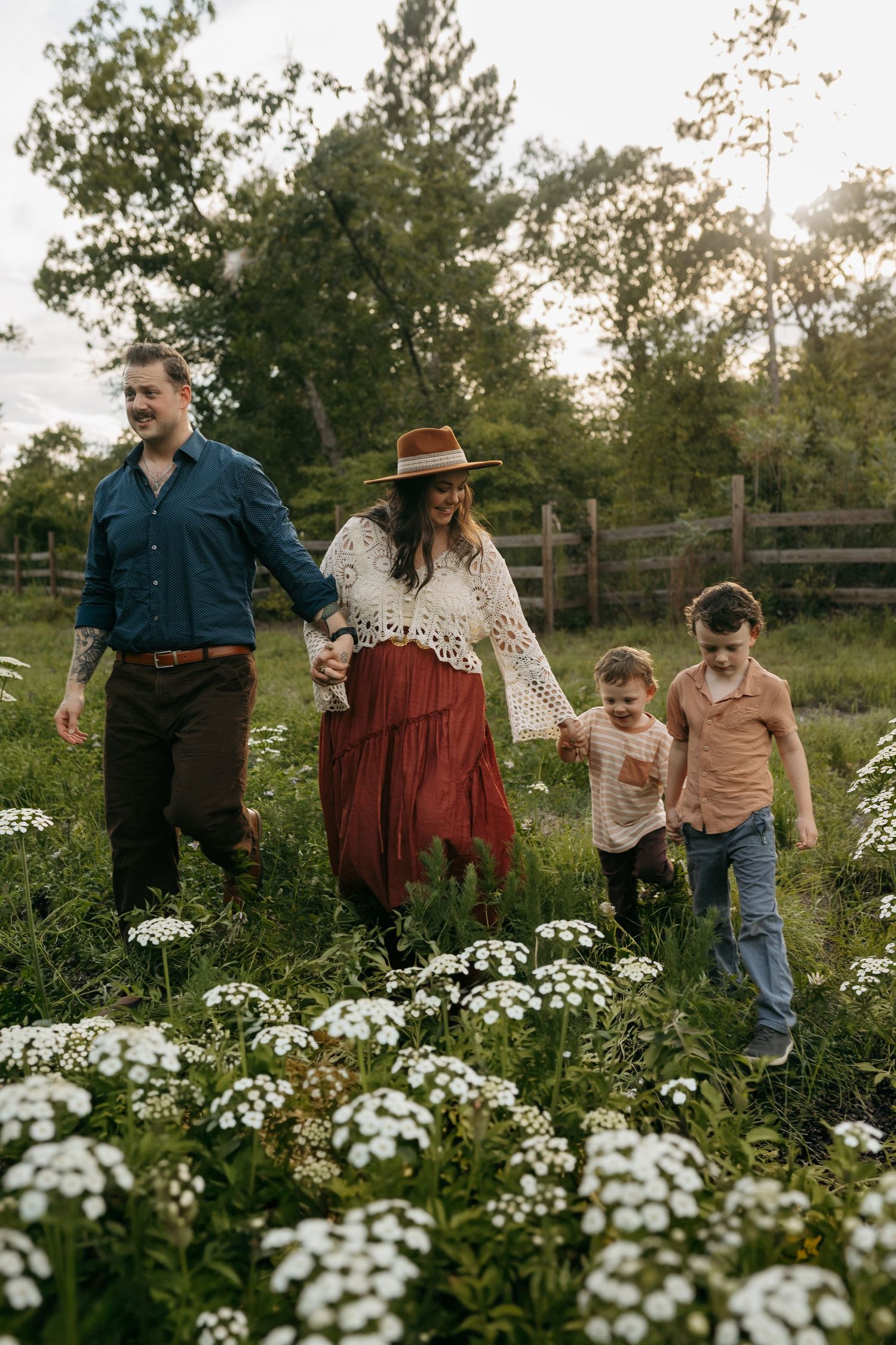 Family of four walks hand-in-hand through a wildflower meadow at golden hour near a wooden fence.