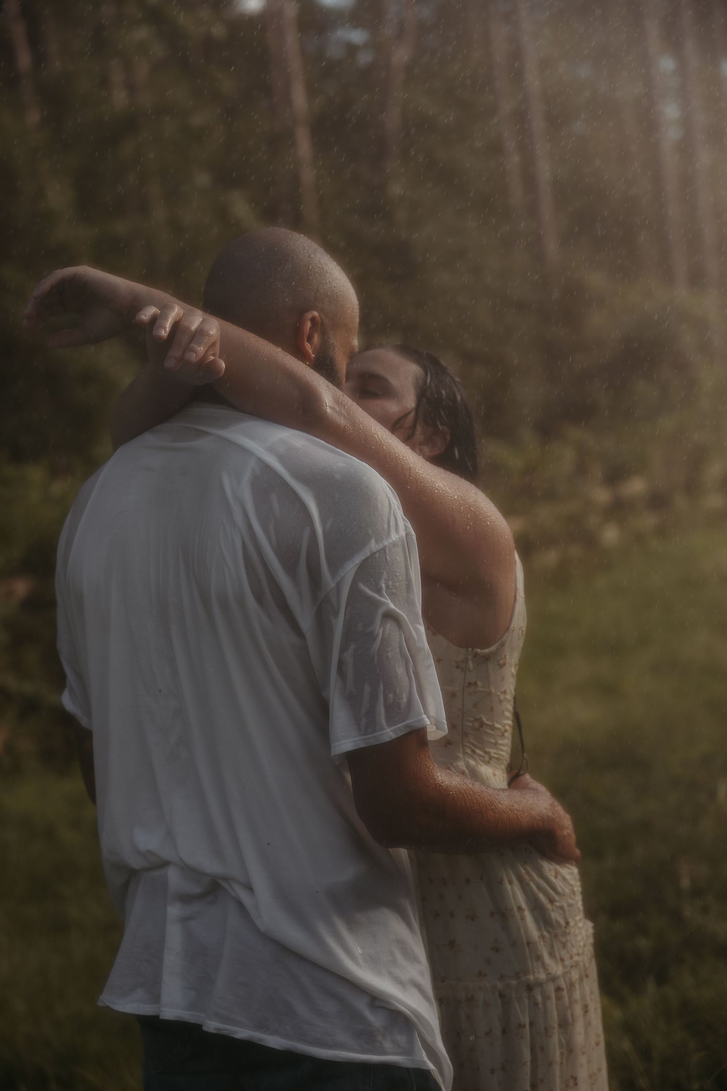 A couple embraces tenderly in a romantic outdoor setting with a dreamy forest backdrop at sunset.