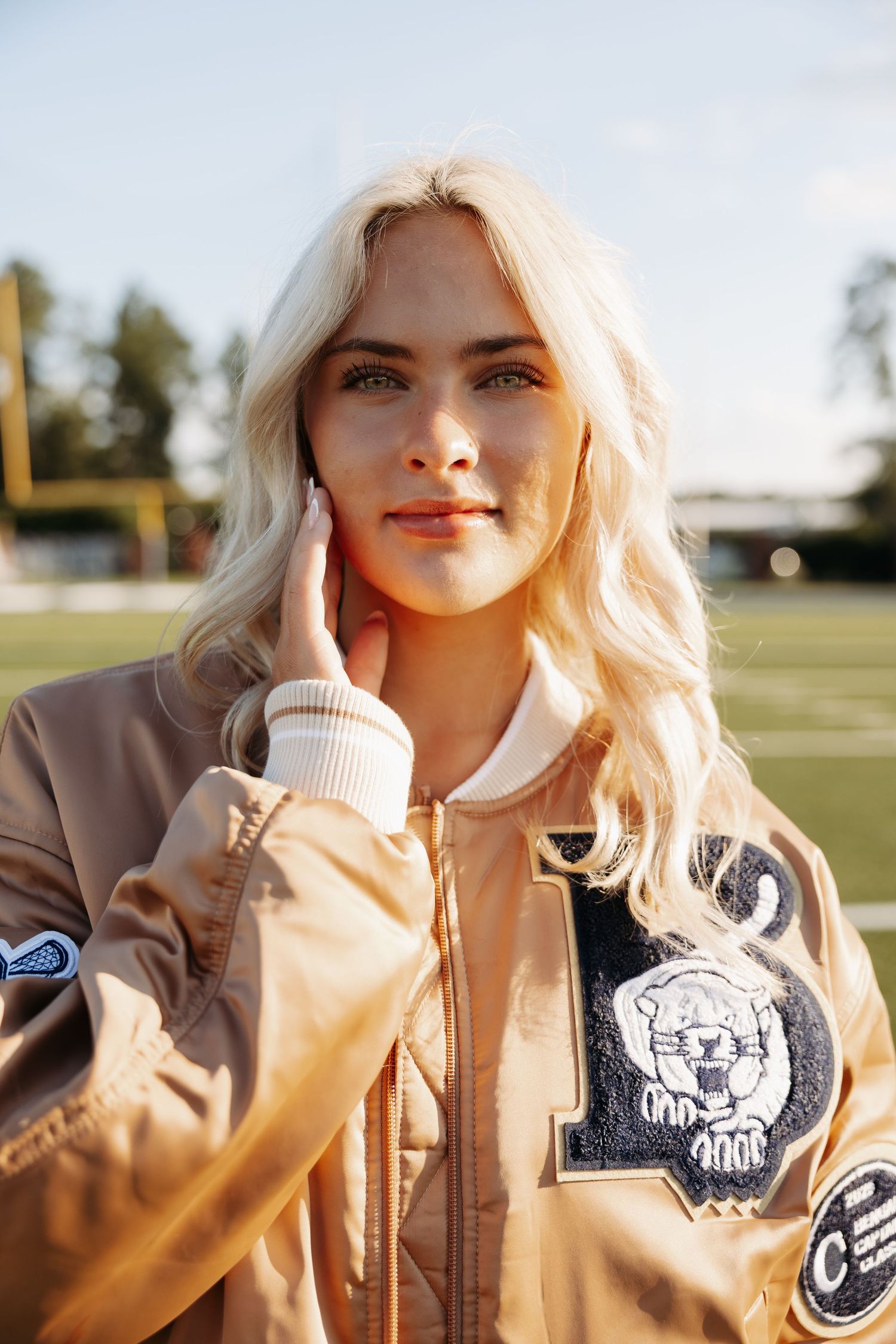 Woman wearing beige bomber jacket with patches poses outdoors in natural lighting.