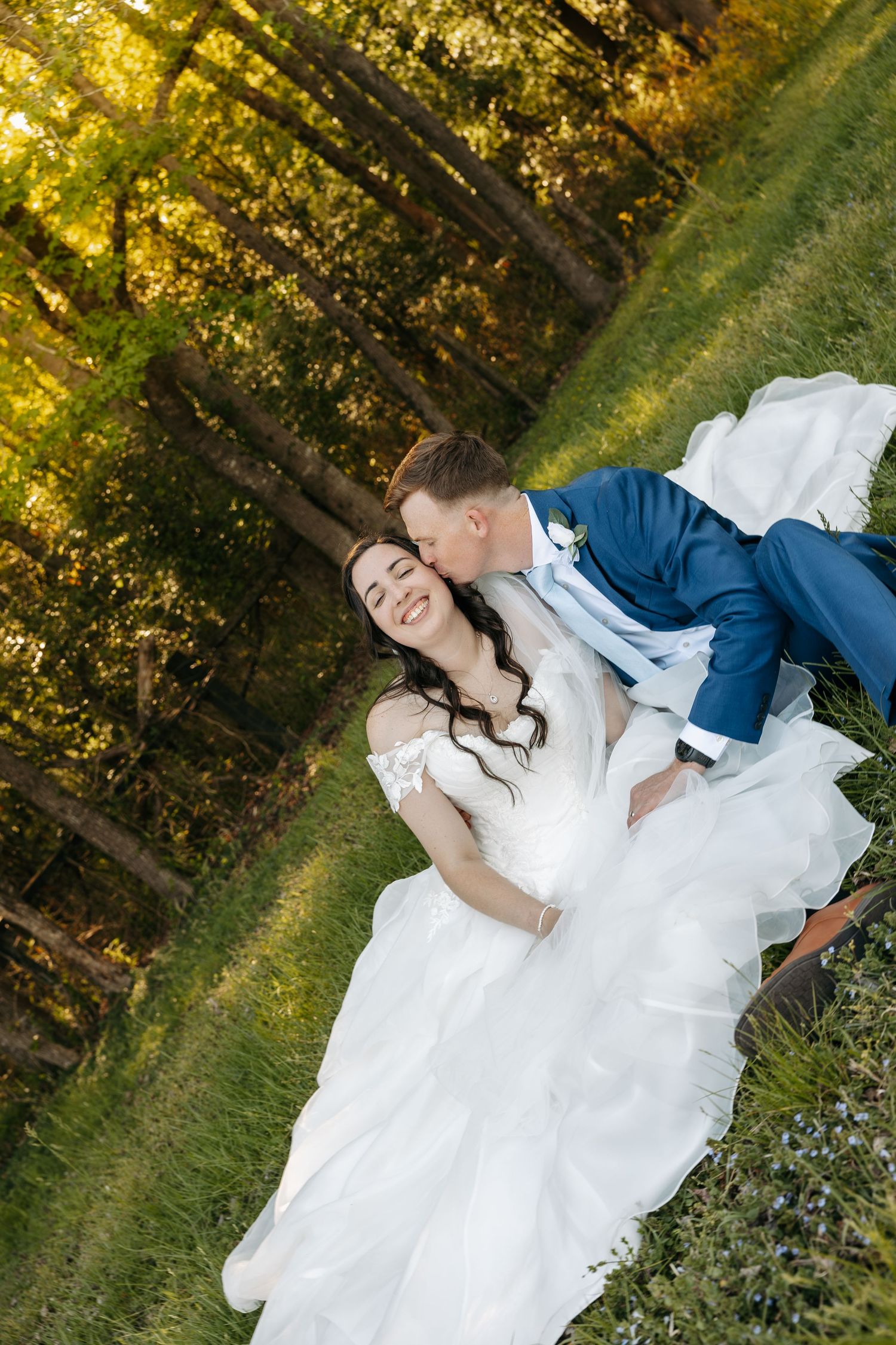 Groom in blue suit kisses smiling bride in white gown as they recline on green grass.