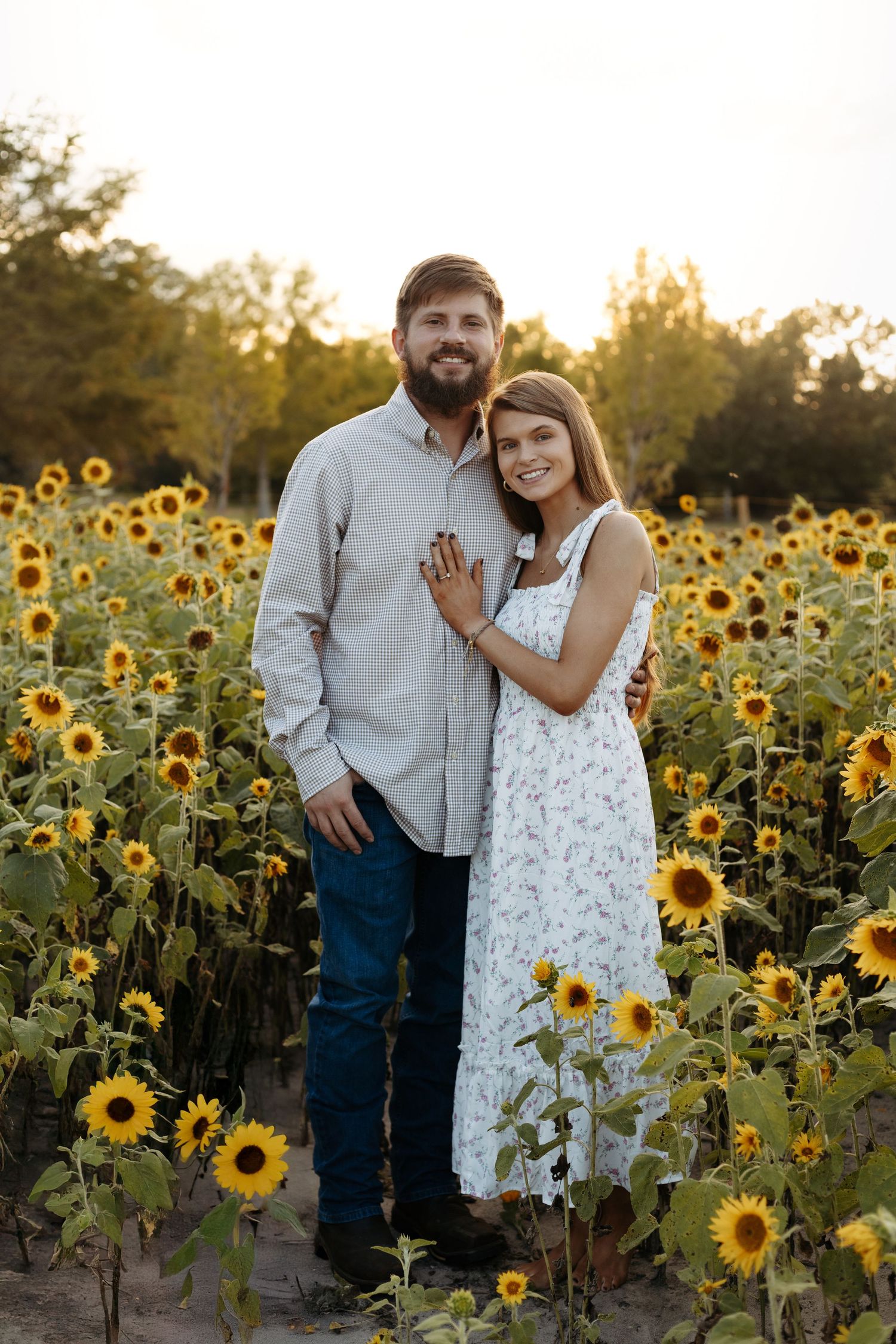 A smiling couple embraces in a sunflower field at golden hour, woman in floral dress, man in blue shirt and jeans.