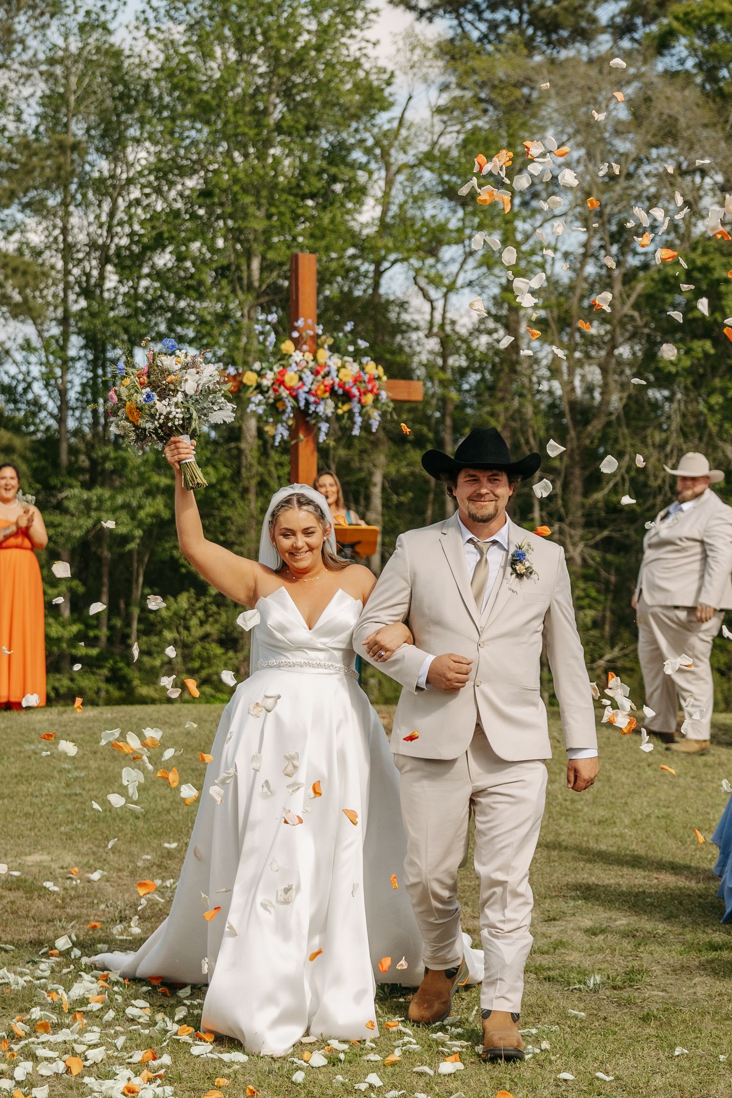 Bride and groom walk down outdoor aisle after wedding ceremony, with flower petals tossed in the air.