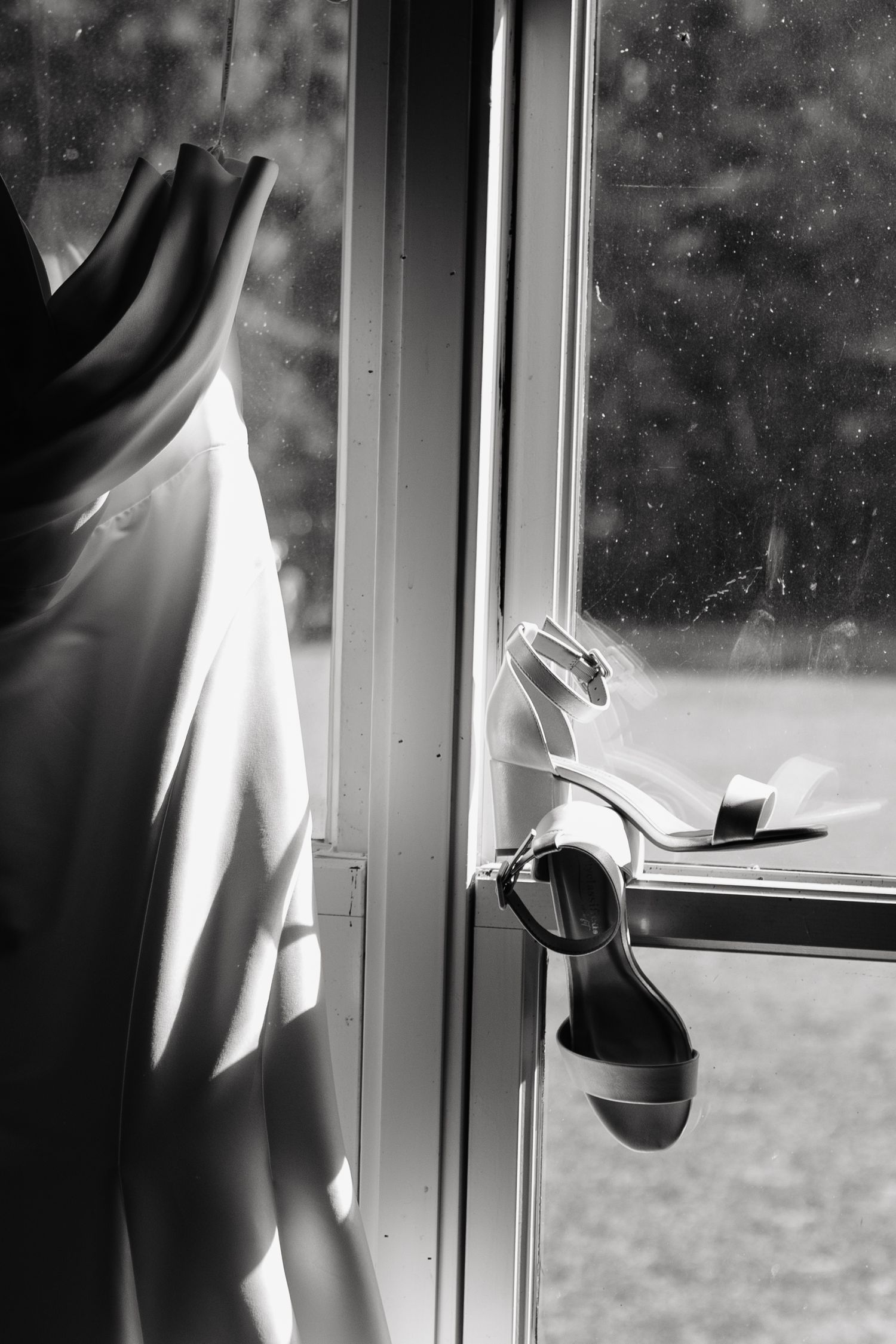 Black and white photo of a wedding dress hanging by a window with bridal heels resting on the window handle.