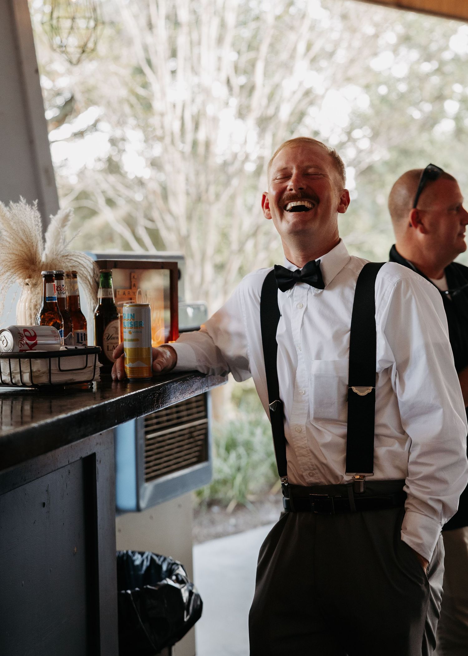 A laughing groom in suspenders and bow tie stands at a bar during a wedding celebration.