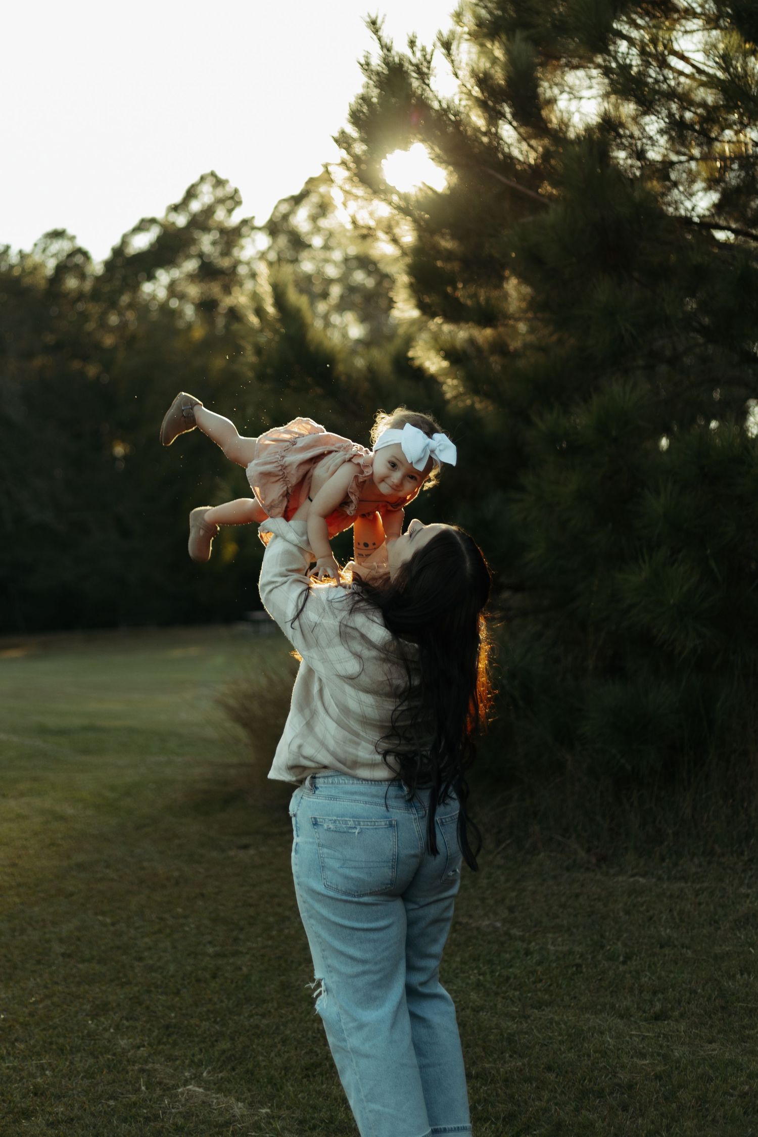 A mother joyfully lifts her baby girl in the air outdoors at sunset, with lush green trees in the background.