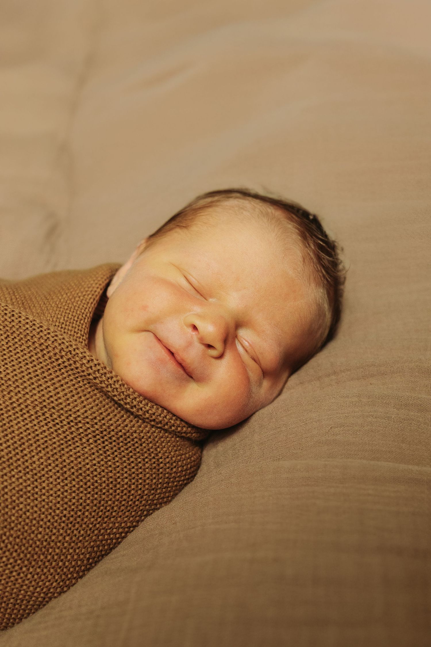 A sleeping newborn swaddled in a soft brown knit wrap rests peacefully on beige bedding.