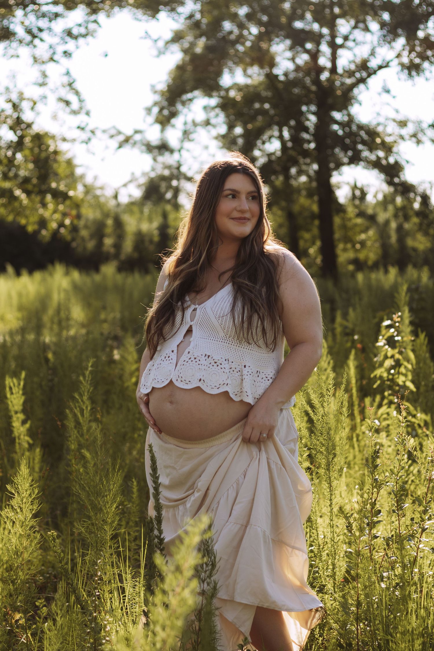 A figure poses in a sunlit meadow wearing a white eyelet crop top and flowing skirt among tall summer grasses at sunset.