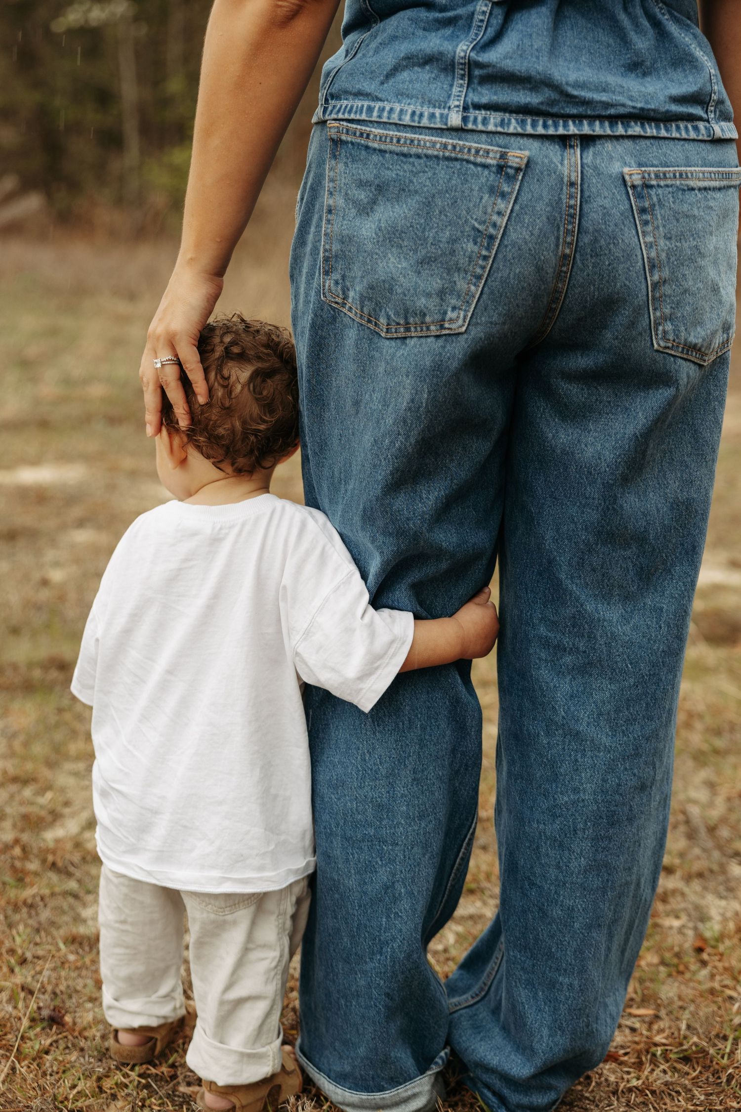 A toddler in white clothing hugs their mother's leg as she stands in jeans outdoors in a natural setting.
