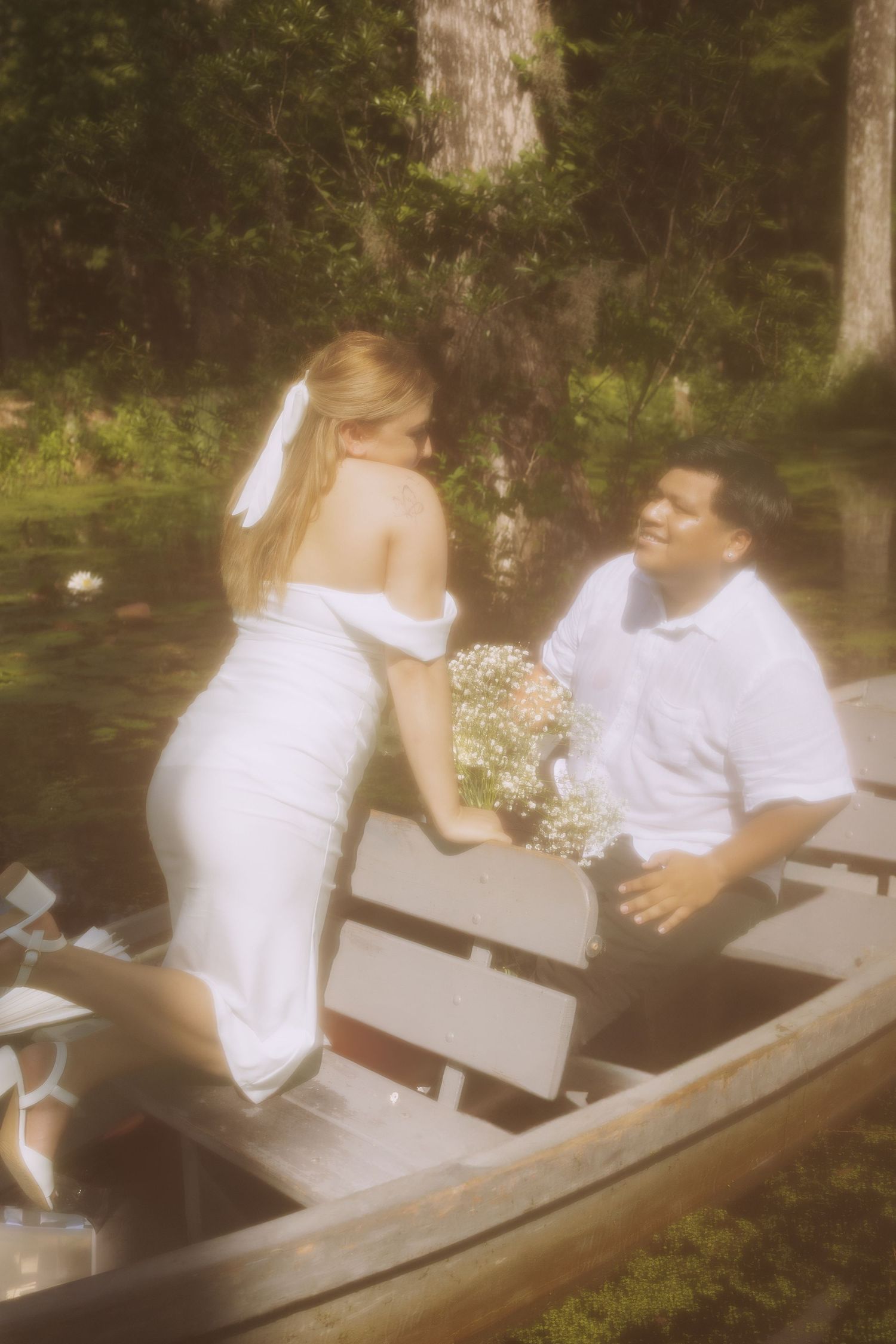 Bride in white dress holding bouquet sits with groom in a wooden canoe surrounded by lush greenery.
