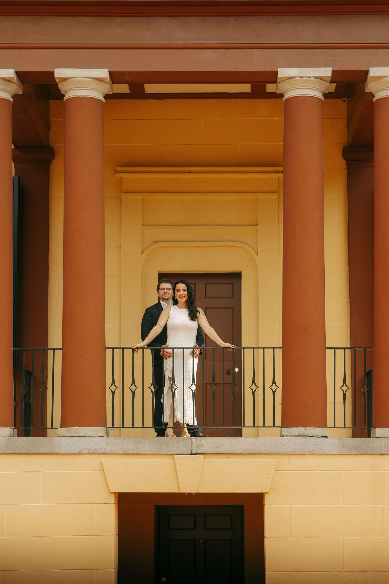 Couple posing on a balcony with tall orange columns and a dark wooden door in the background.