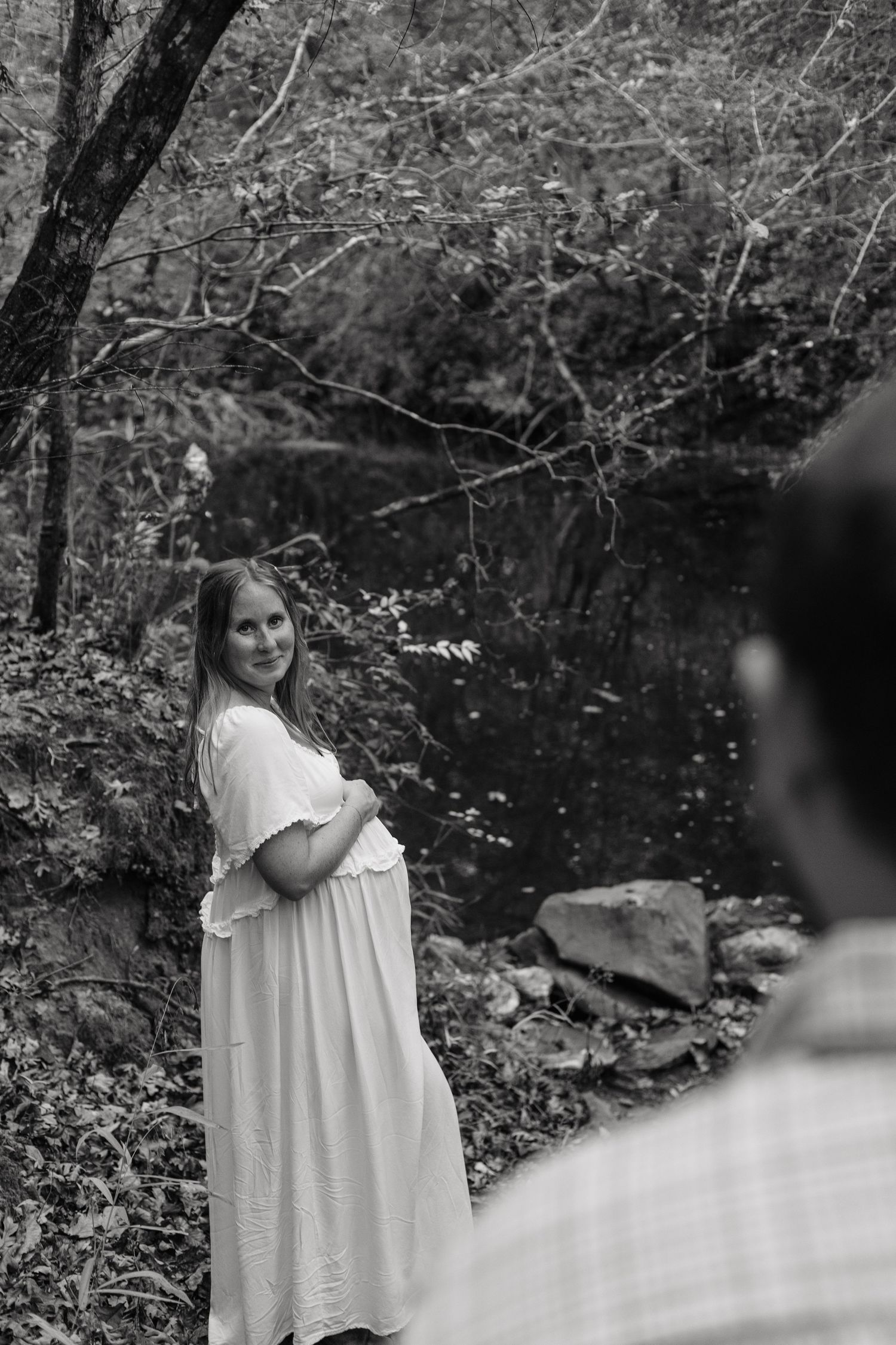 Black and white photo of a pregnant woman in a white dress smiling in a lush forest setting near a rocky stream.