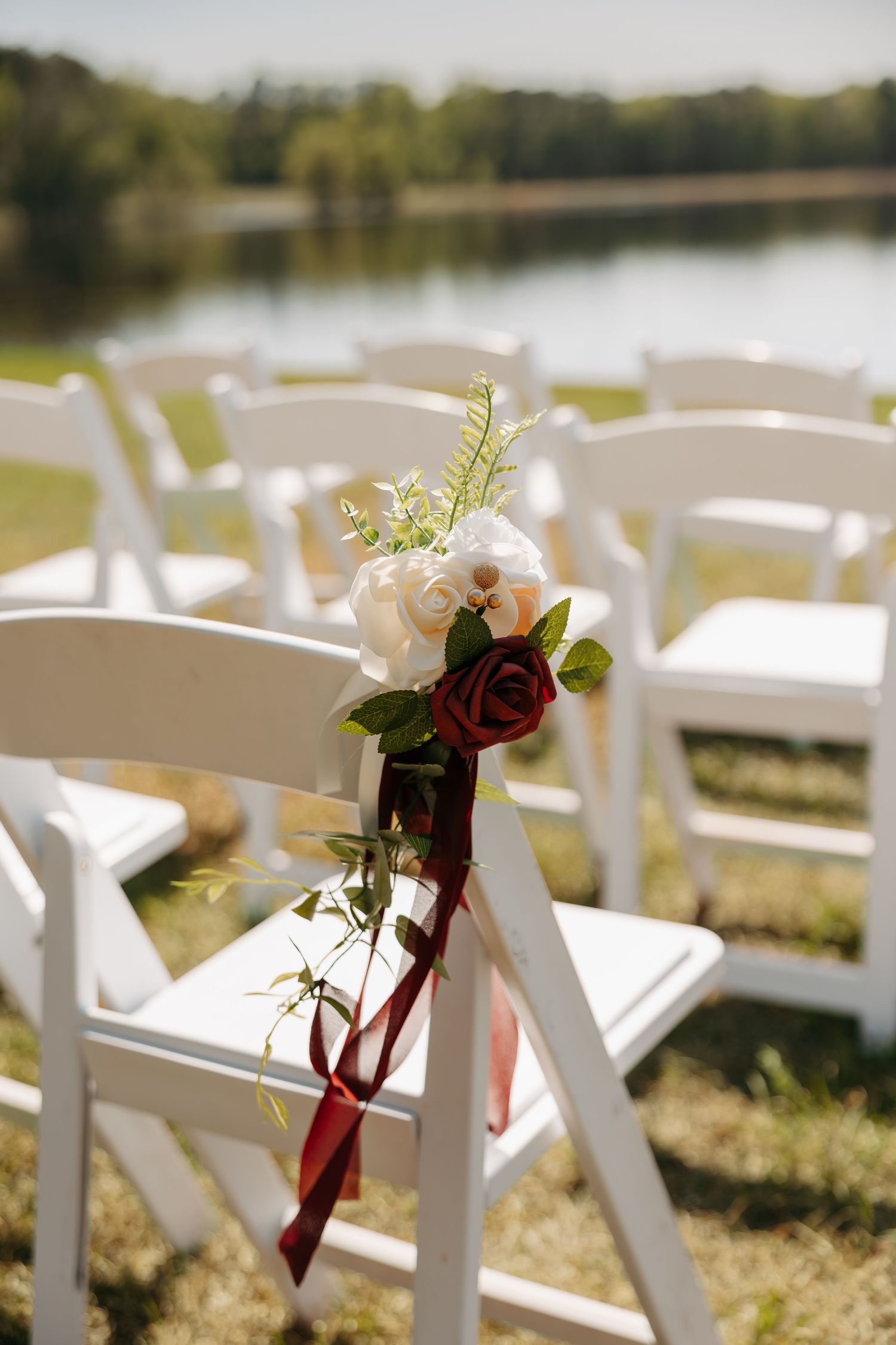 White wedding chairs adorned with red and white roses tied with burgundy ribbon at an outdoor lakeside ceremony.