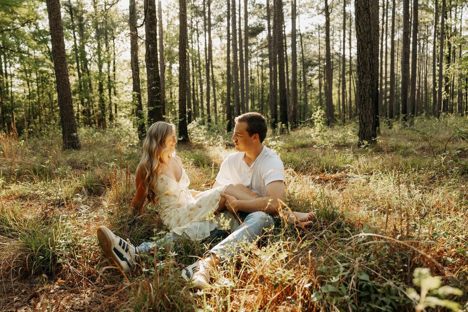 Couple sitting together on forest floor surrounded by tall pine trees in warm golden sunlight.